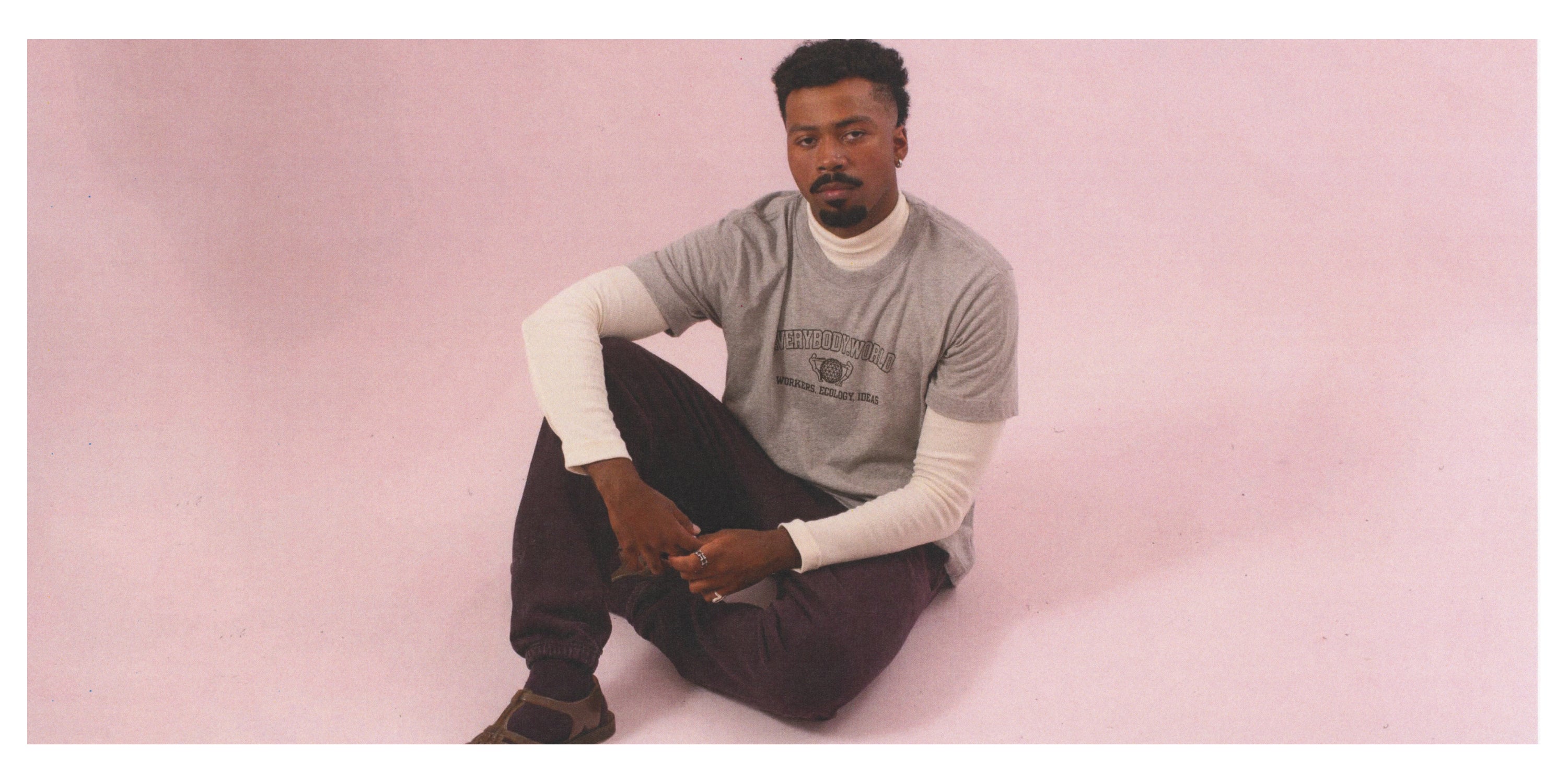 Man sitting on a pink background wearing a Heather Short Sleeve Tee that reads 'Everybody.World' and Trash Sweatpants.