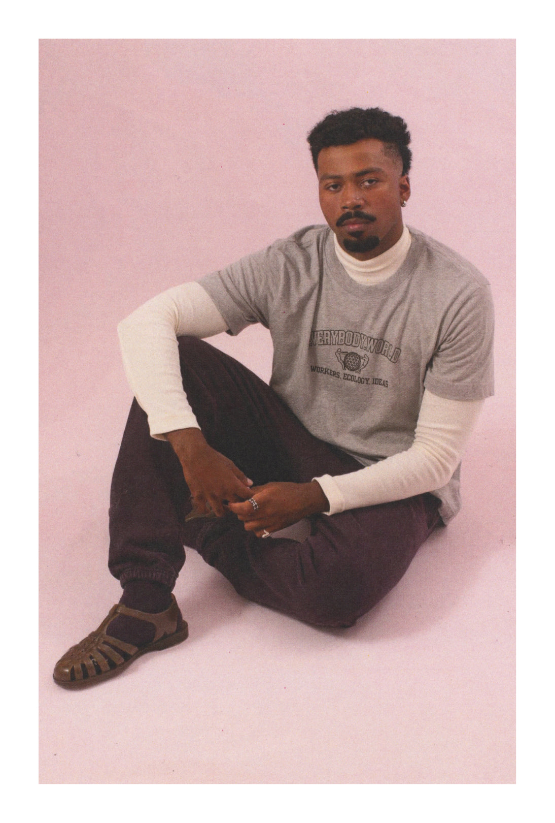 Man sitting on a pink background wearing a Heather Short Sleeve Tee that reads 'Everybody.World' and Trash Sweatpants.