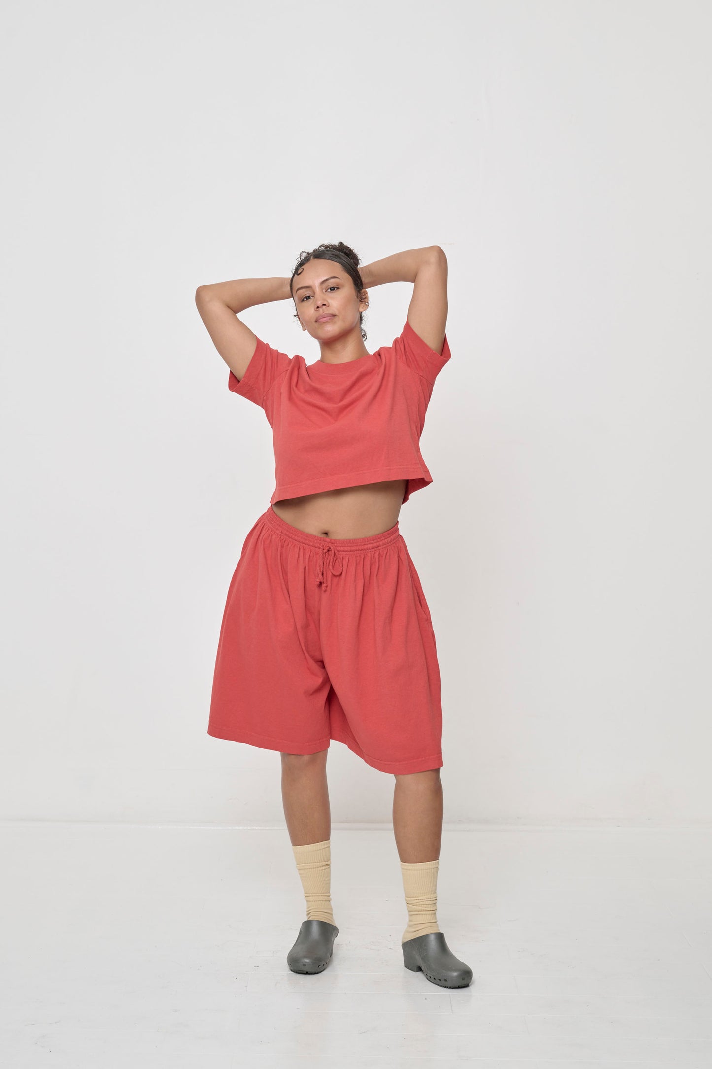 Styled with matching shorts, hands behind her head raises shirt hem to expose midriff.