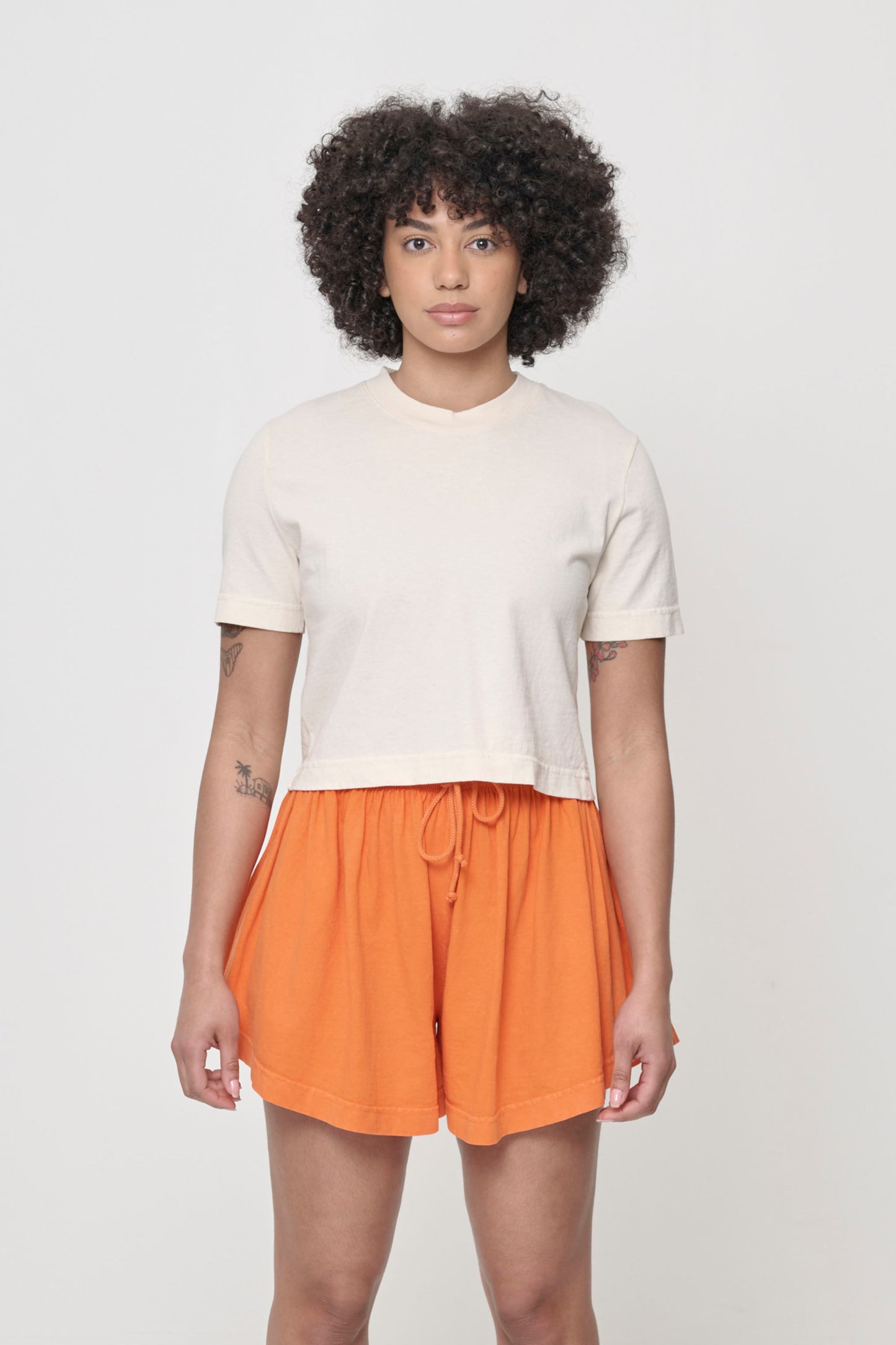 A woman wearing cropped light beige t-shirt and bottom hem hitting bottom of shorts waistband.
