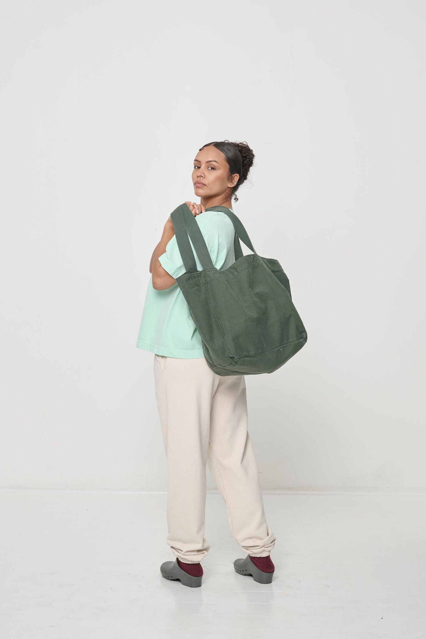 Woman holding Big Tote in Pine greyish dark green over her shoulder, tilting the bag up to show the deep bottom panel.