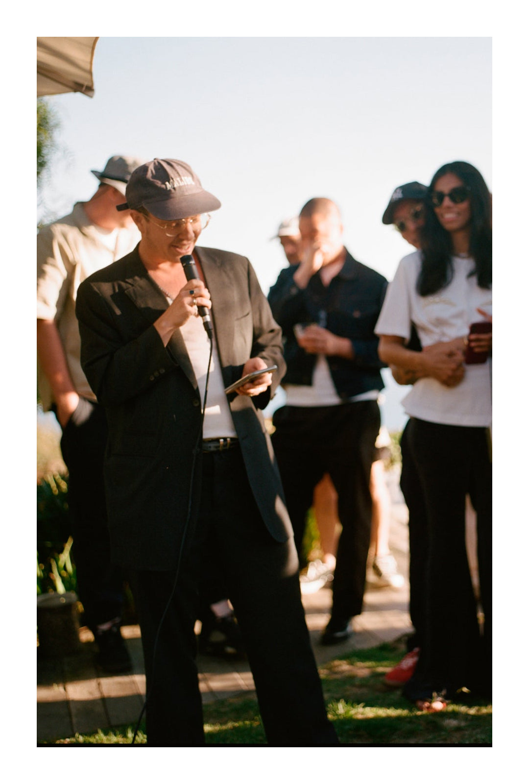 Ariel Katz, in a black suit, speaks into a microphone, holding a phone, at an outdoor event with a small crowd.