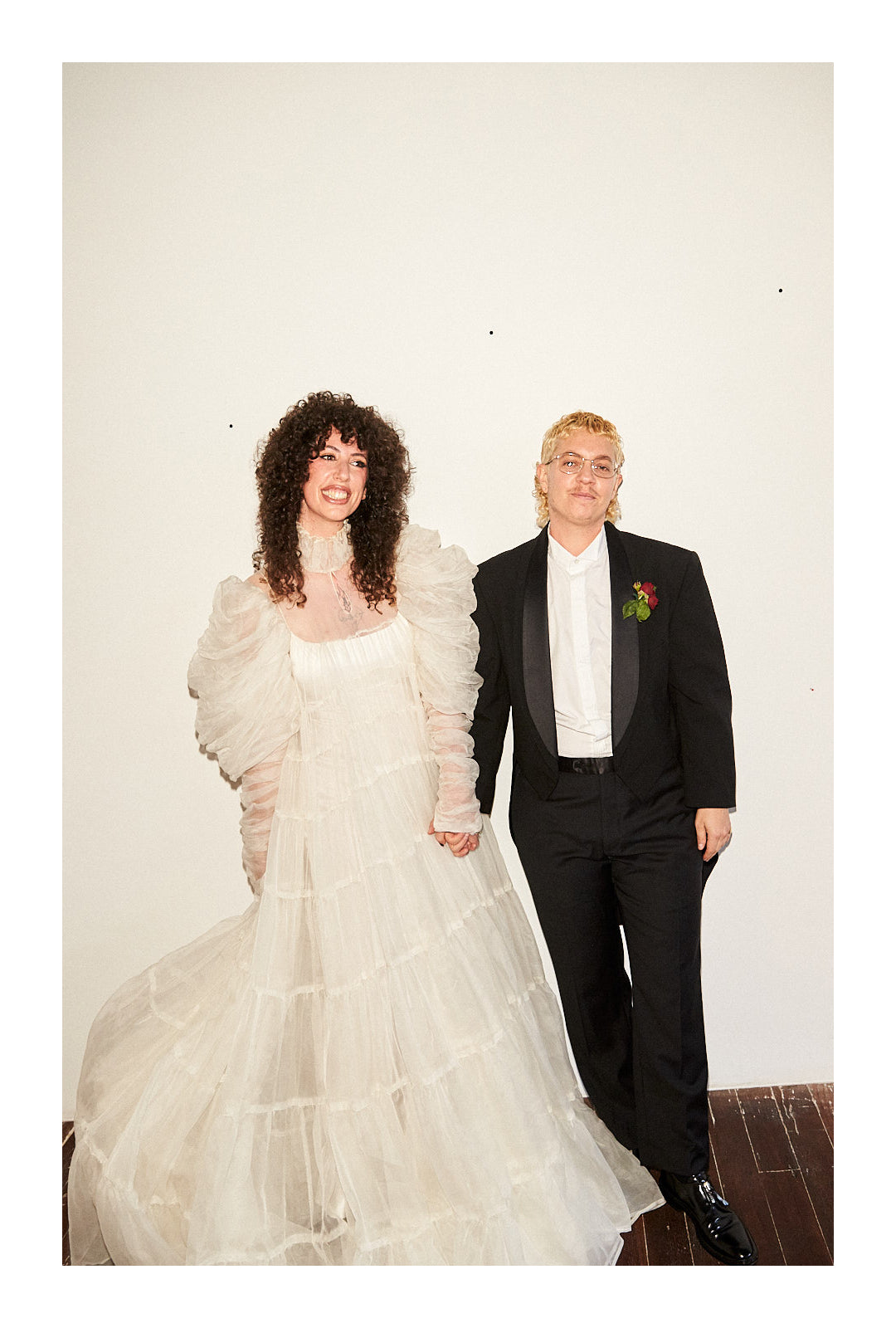 Jerico in wedding dress and Ariel in a tux, holding hands in front of a white wall.