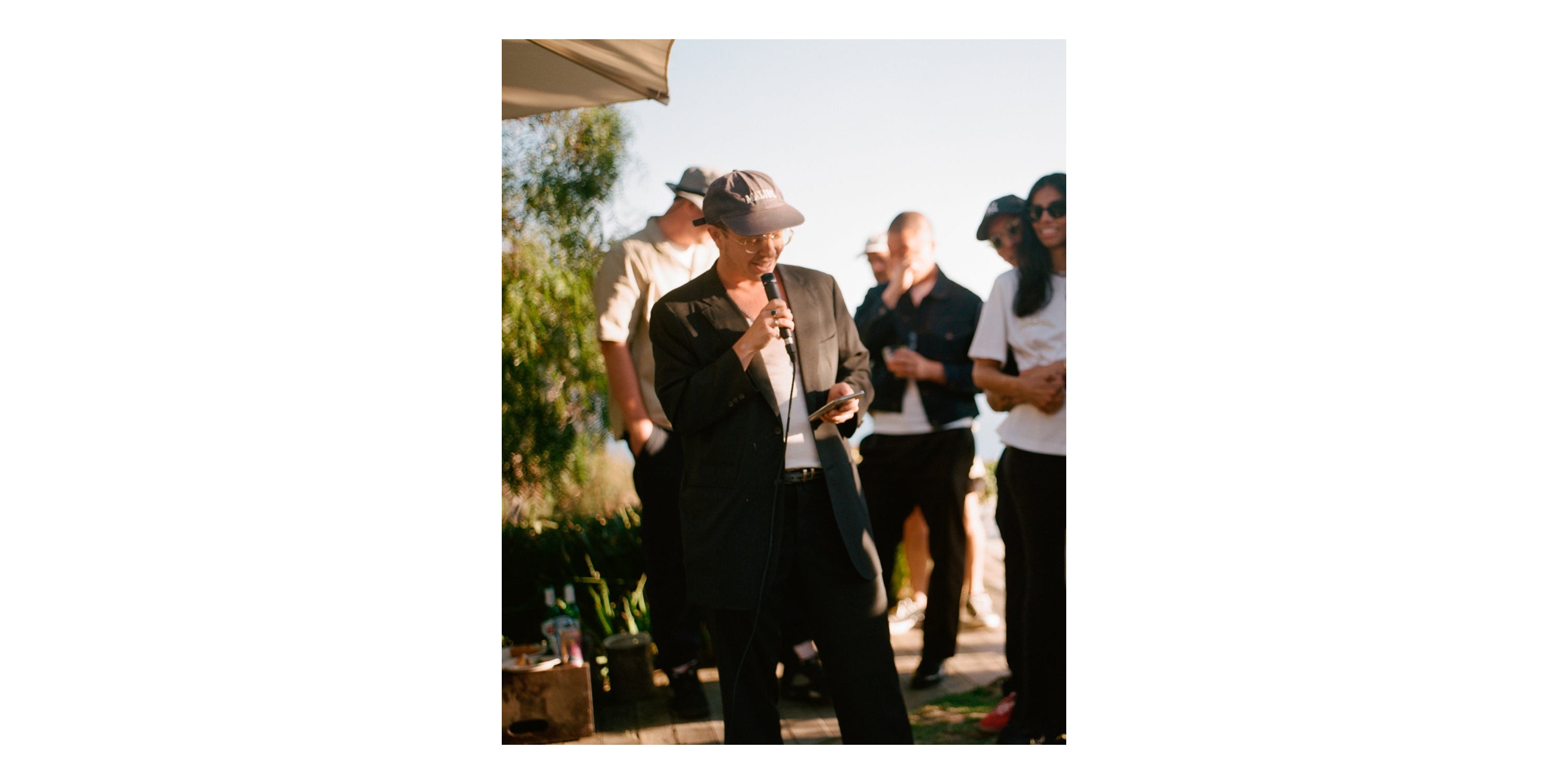 Ariel Katz, in a black suit, speaks into a microphone, holding a phone, at an outdoor event with a small crowd.