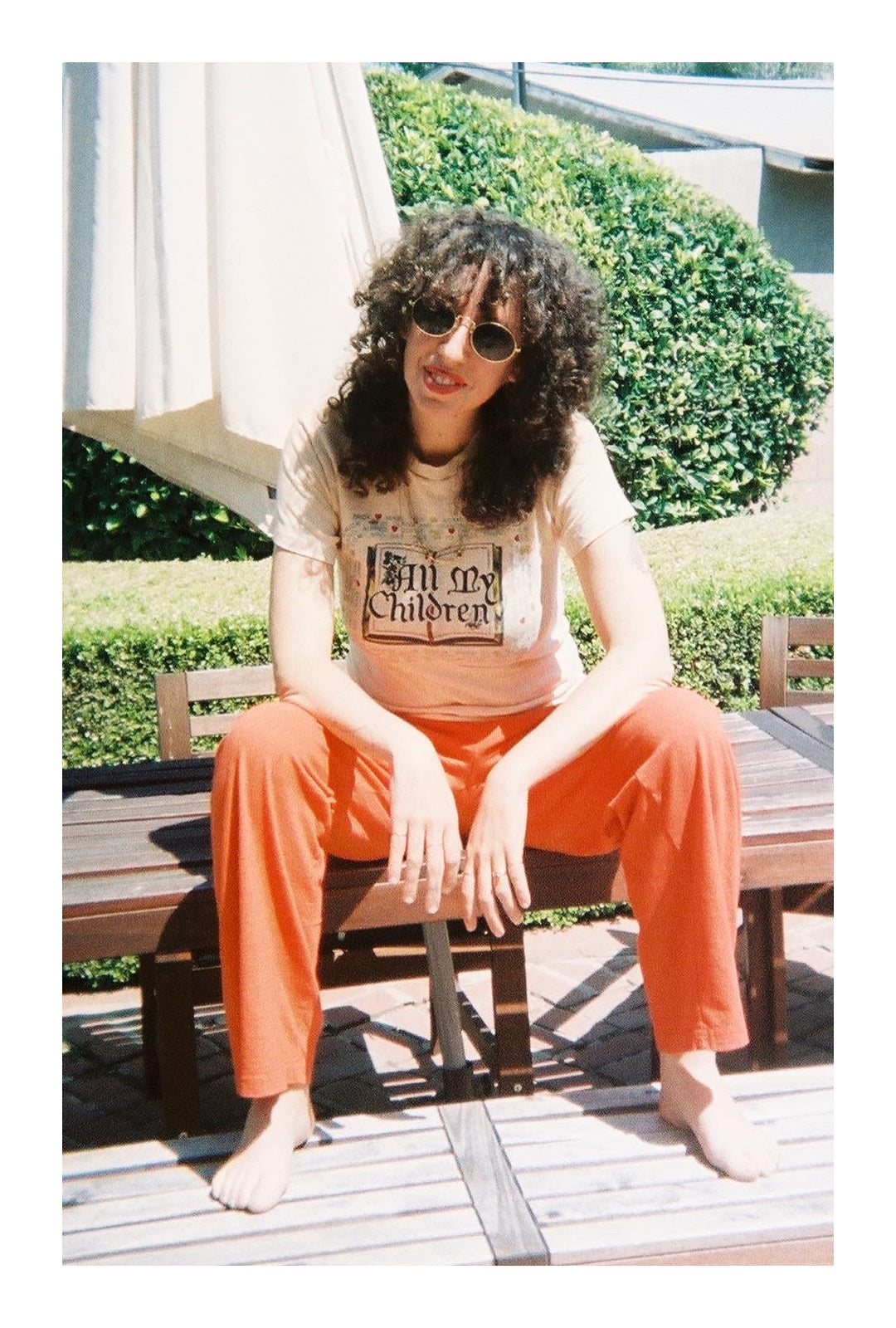 Jerico in an "All My Children" tee and Orange/Red Apartment Pants, sitting on a wooden picnic table.