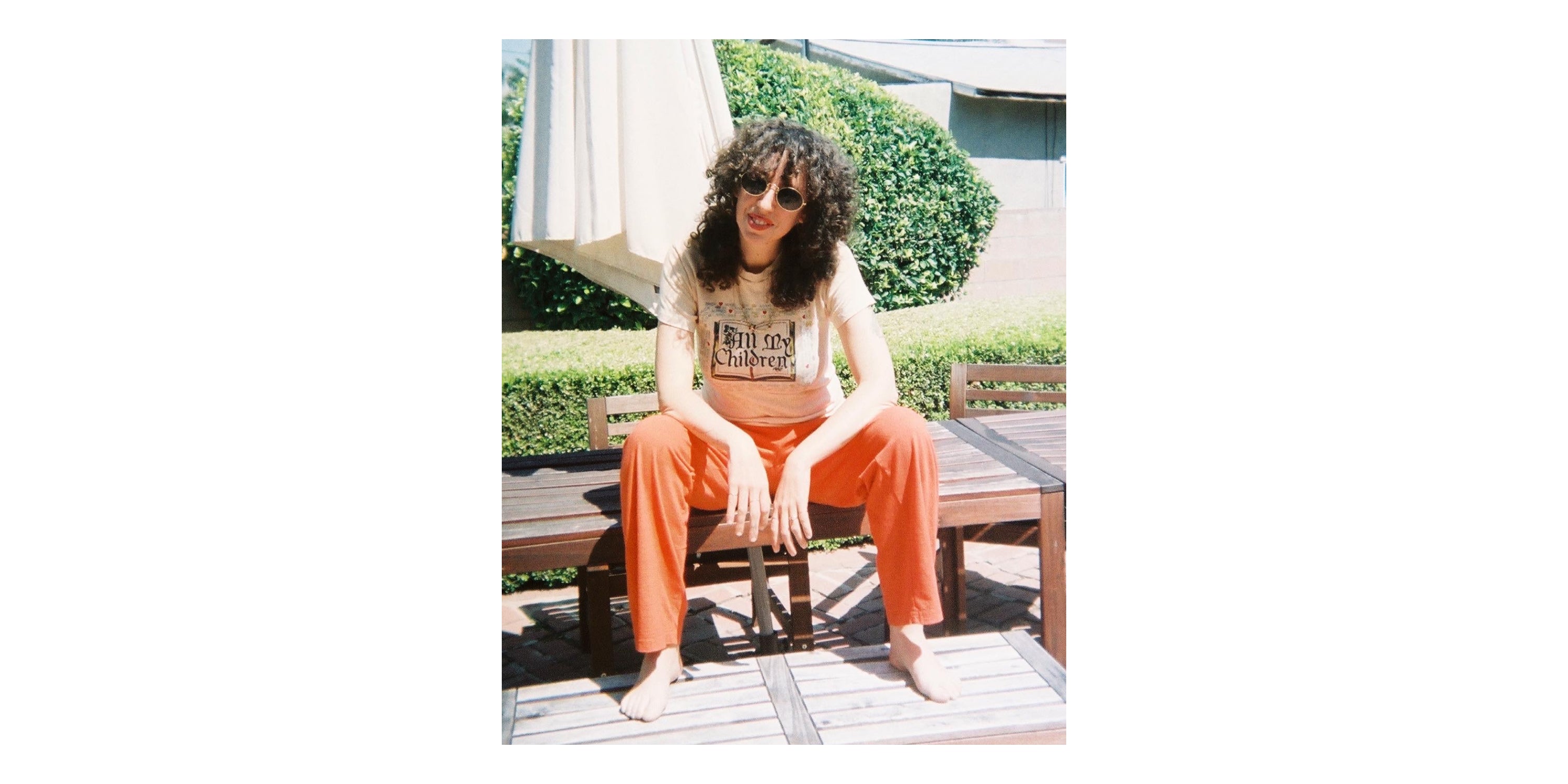 Jerico in an "All My Children" tee and Orange/Red Apartment Pants, sitting on a wooden picnic table.
