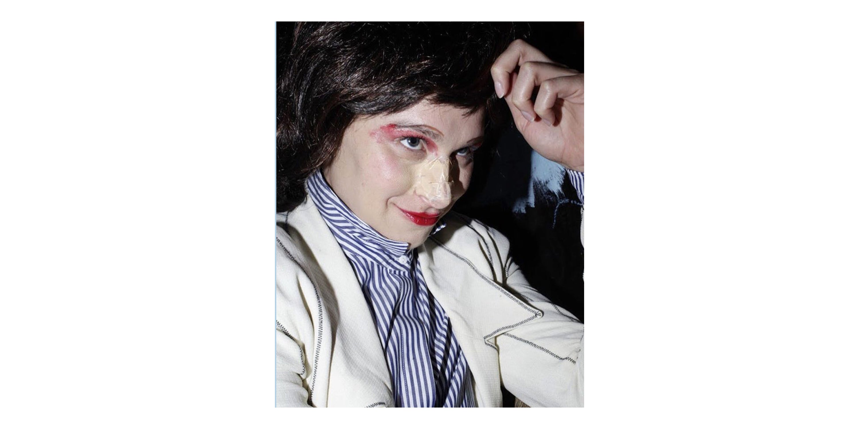 Close up of Mae in dark brown wig, striped button up shirt, and off white jacket. Red lipstick on lips and eyelids, and tape on her nose.