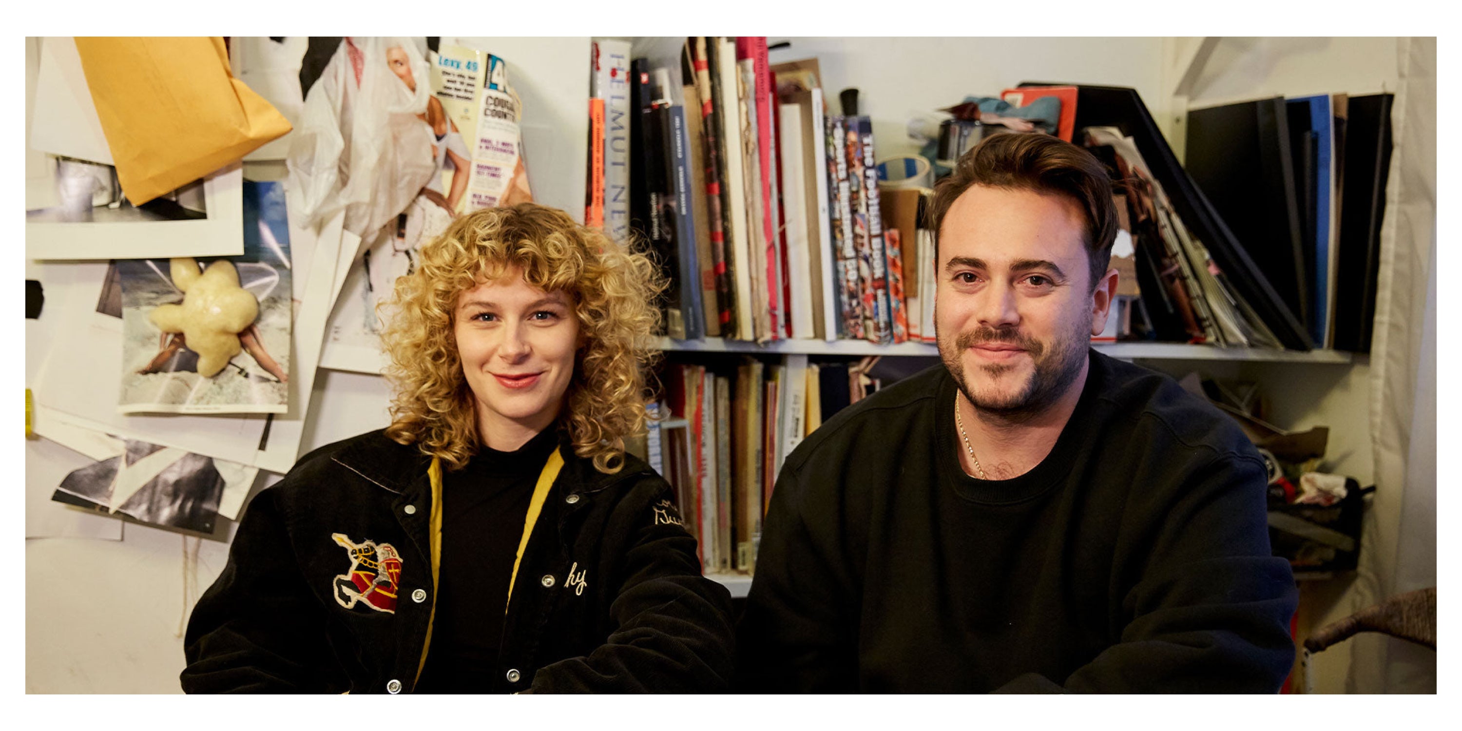 Mae and Kalen sit next to each other, smiling. Background are books and magazine clippings attached to the wall.