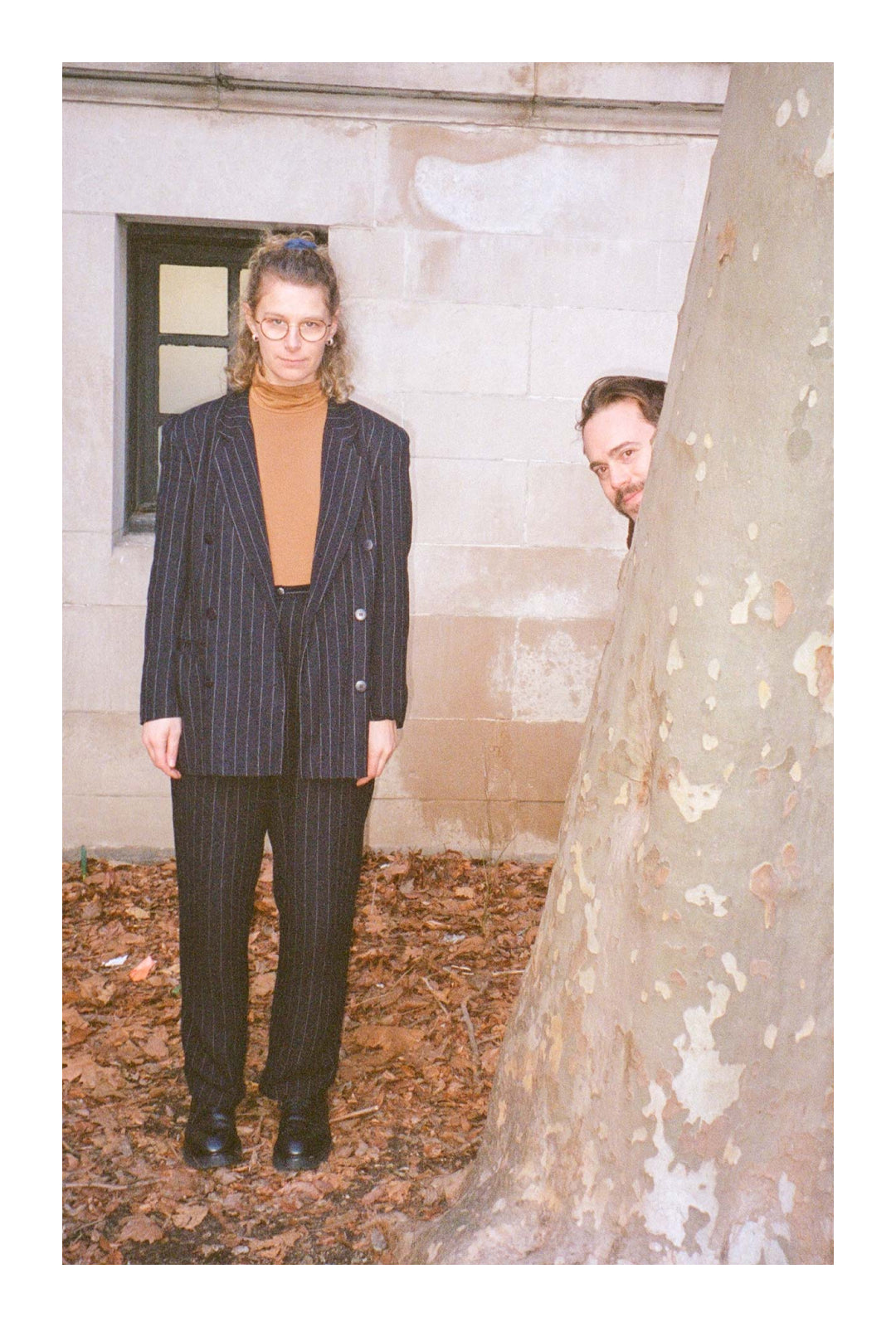 Mae and Kalen in front of a white brick building with a black window and dried leaves. Kalen peeks from behind a tree; Mae, next to the tree, wears a pinstripe suit and an orange JAS Mockneck.