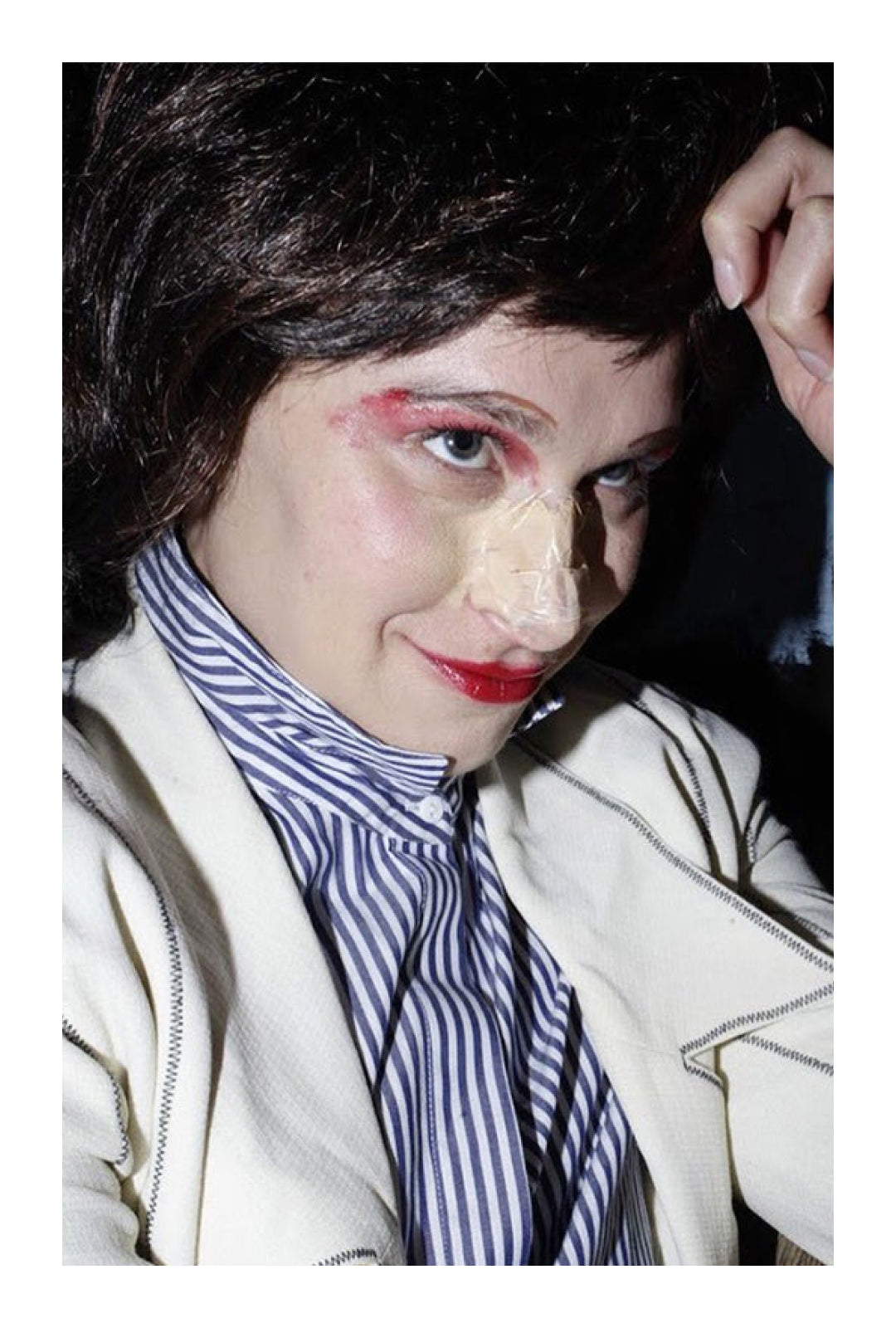 Close up of Mae in dark brown wig, striped button up shirt, and off white jacket. Red lipstick on lips and eyelids, and tape on her nose.
