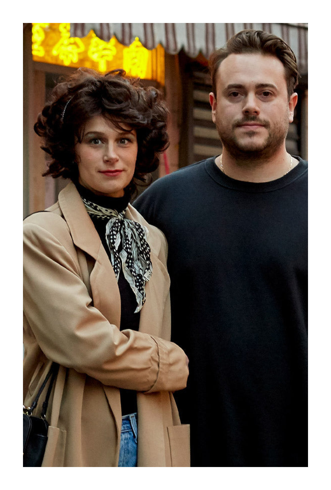 Mae and Kalen stand next to each other, outside in front of a storefront with neon Chinese writing.