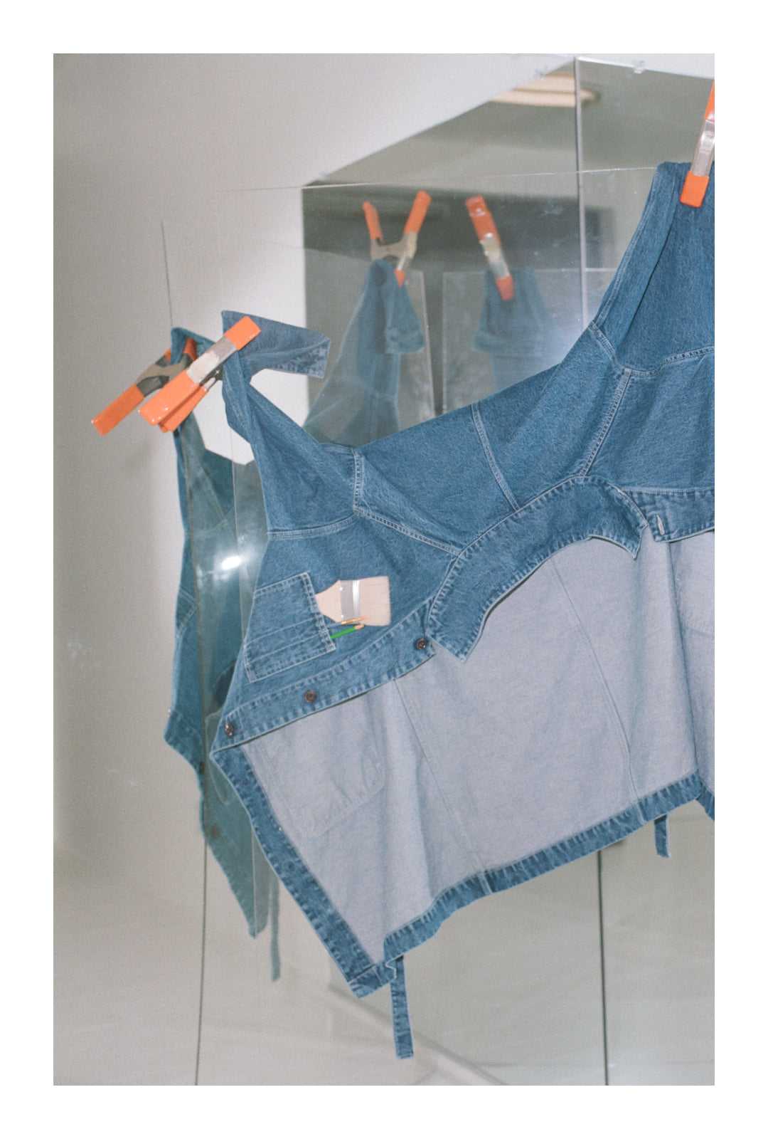 A Denim Griffin Mill Coat clamped to a clear panel, before full-length mirrors. Paintbrushes and pencil in front pocket.