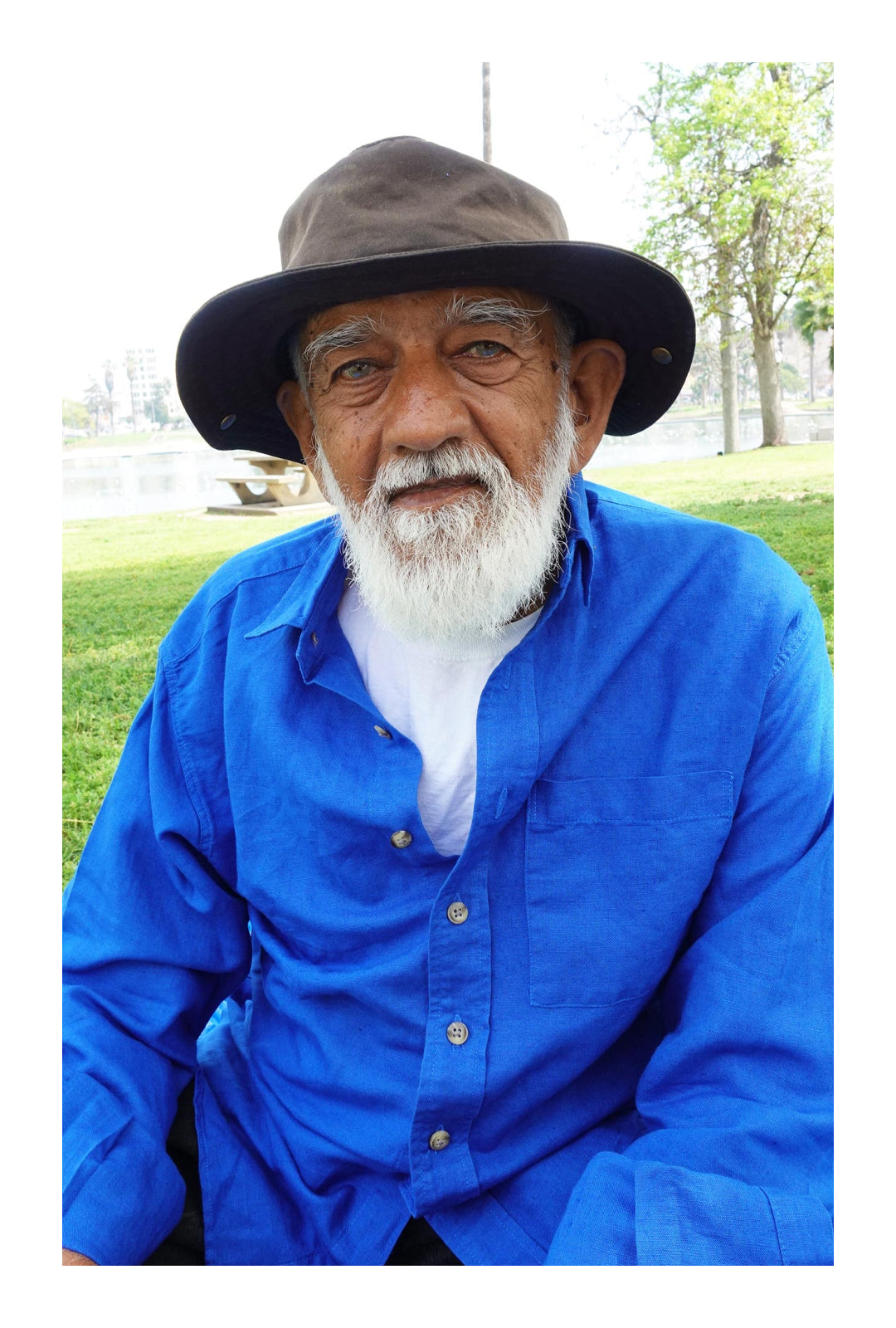 Prakash, in a green sun hat, white beard, and blue shirt, sitting on a park lawn.