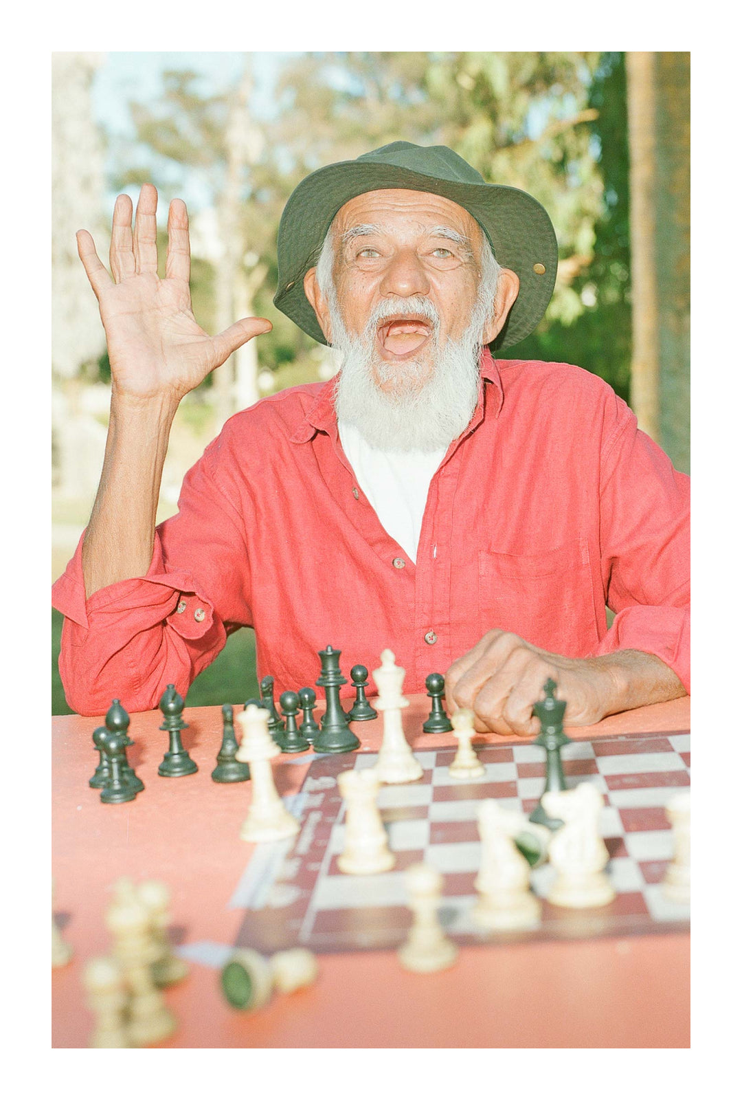 Prakash, in a green sun hat, white beard, and red shirt, gestures hello at a chess table.