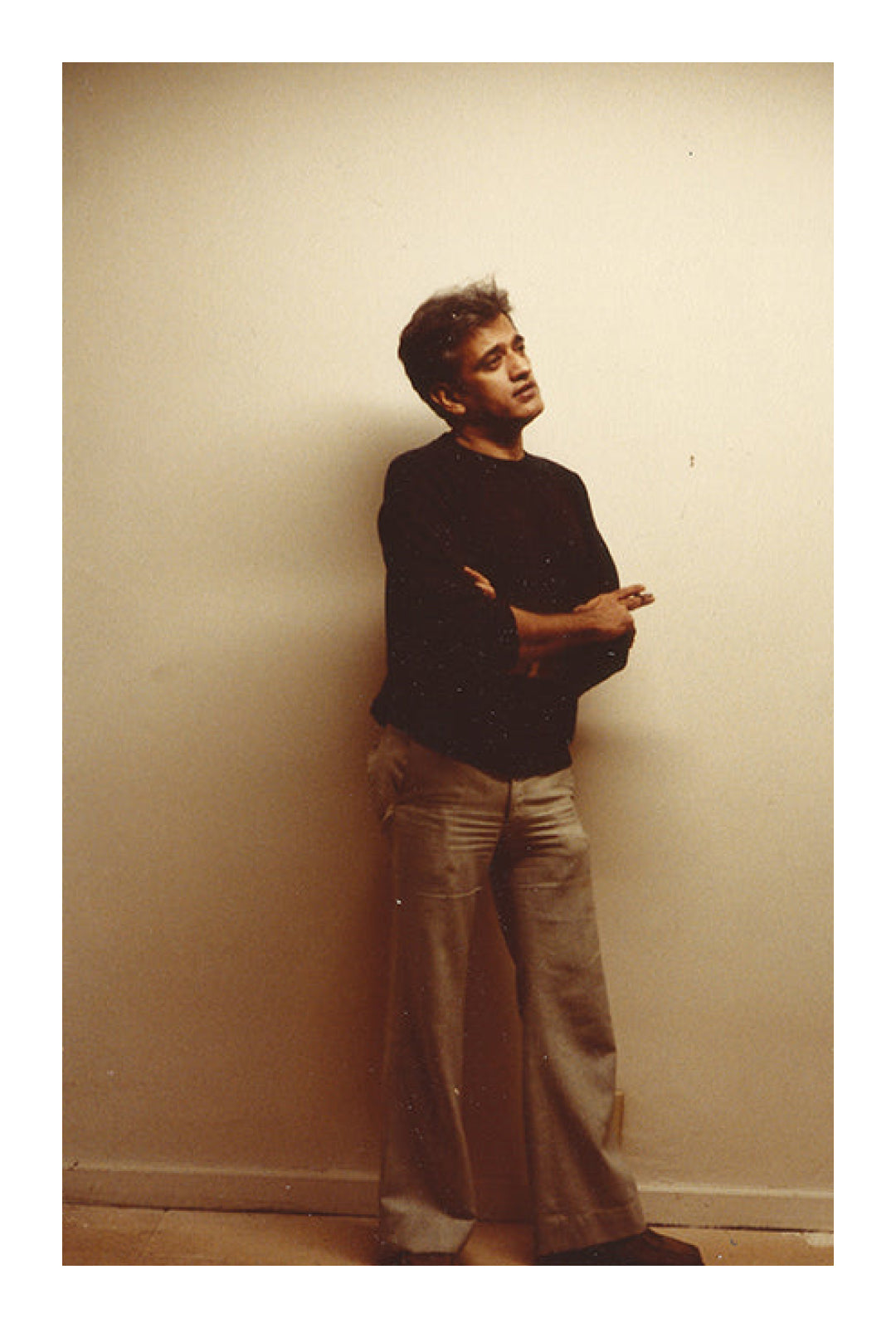 Prakash from decades ago, with short black hair, black sweater, and grey bell-bottom jeans, stands with crossed arms in front of a white wall.