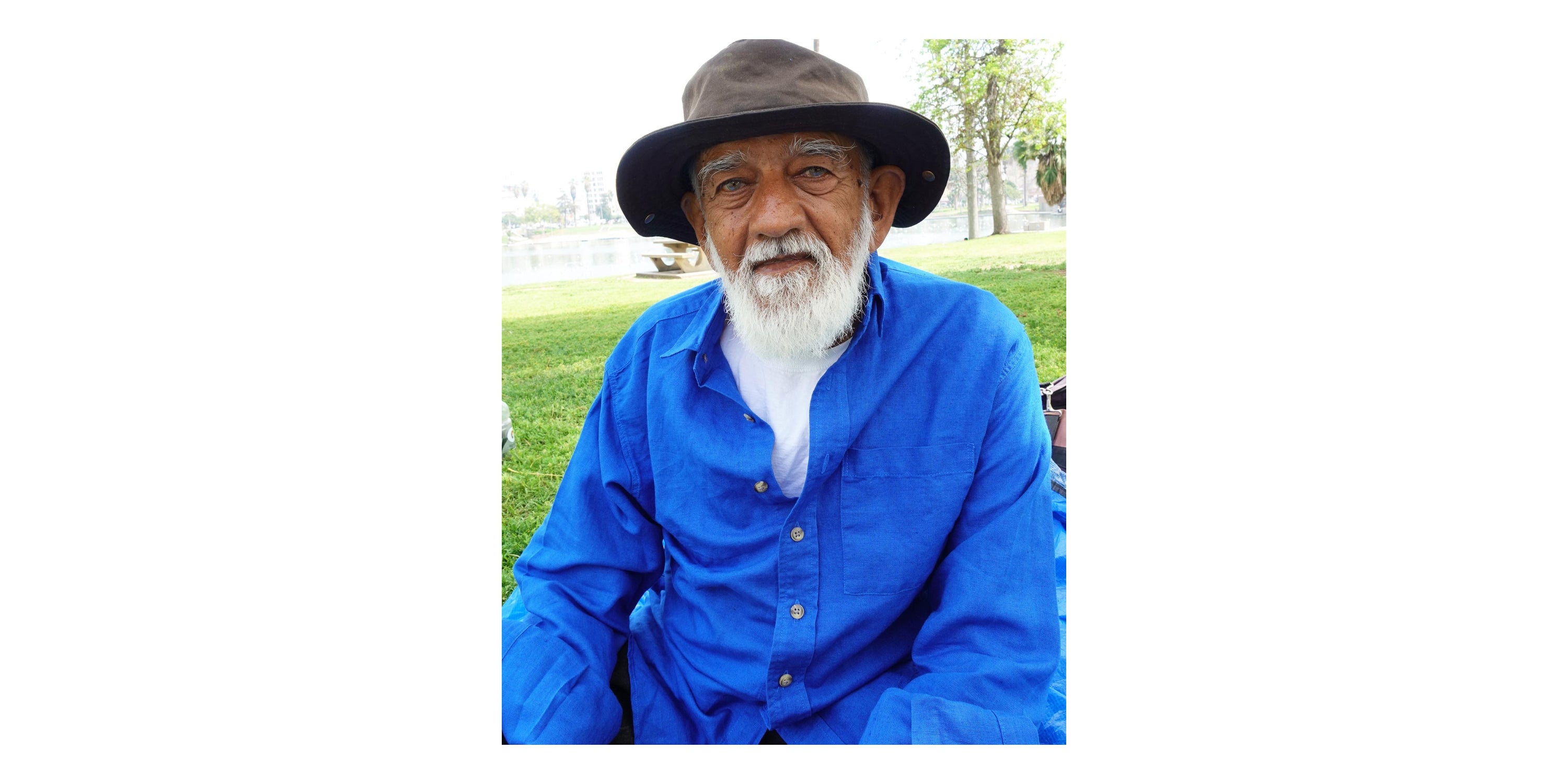 Prakash, in a green sun hat, white beard, and blue shirt, sitting on a park lawn.