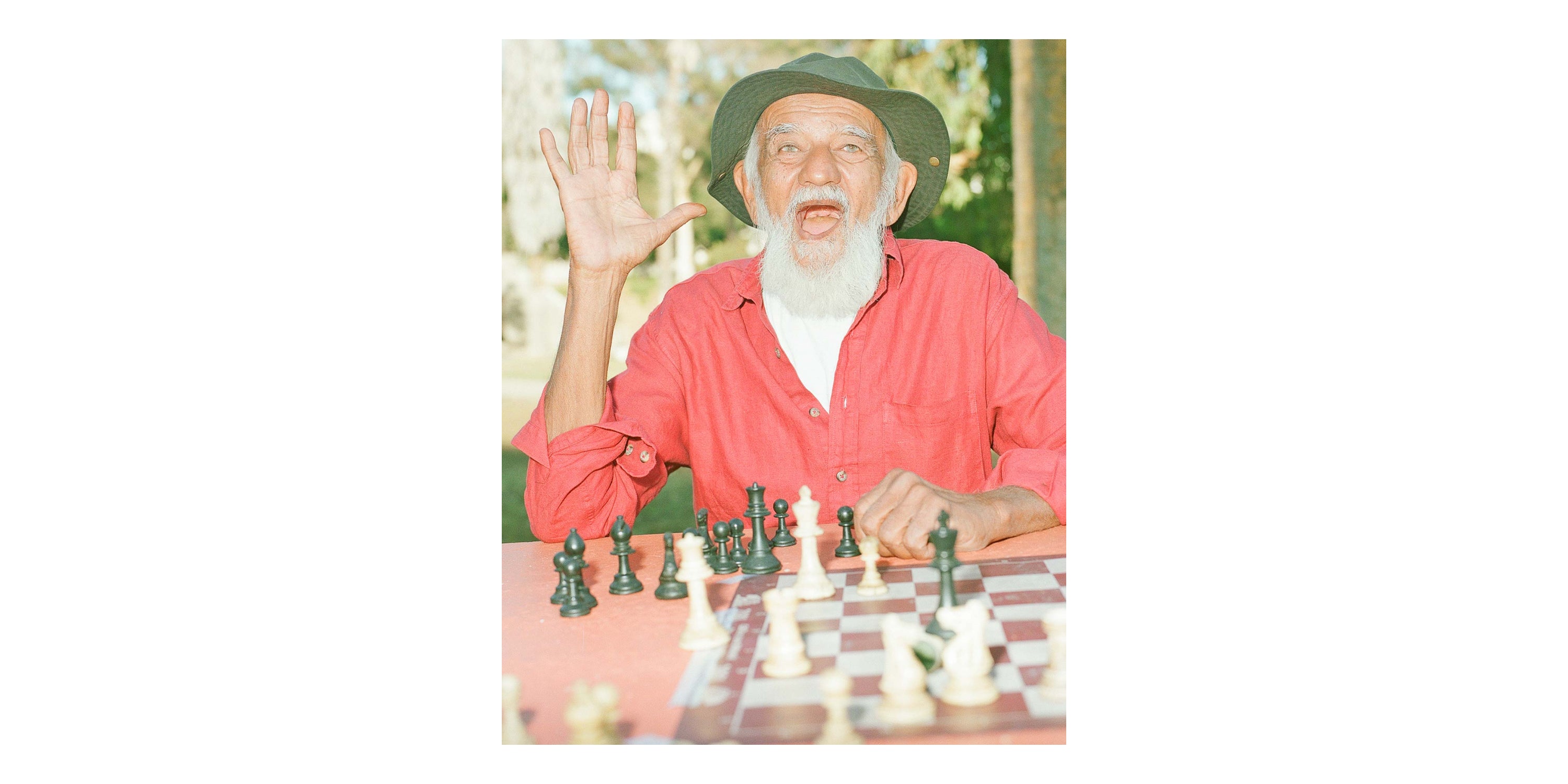 Prakash, in a green sun hat, white beard, and red shirt, gestures hello at a chess table.