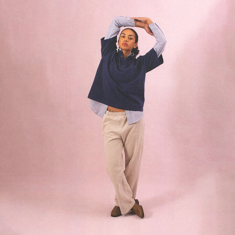 Person stretching arms up, wearing Fleece Apartment Pants in Oat, with Washed Navy Trash Polo over Striped Button-Up.