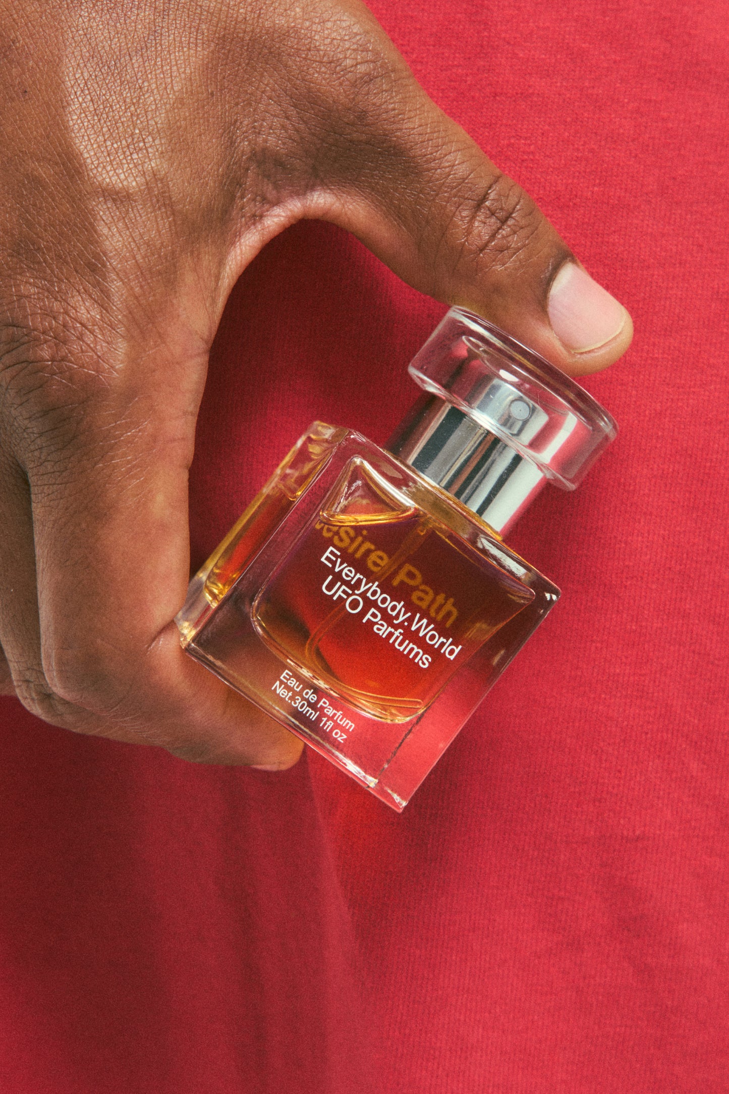 A clear glass bottle of Desire Path Eau de Parfum with a silver cap, containing a yellow-colored liquid, held in a person's hand to show scale.