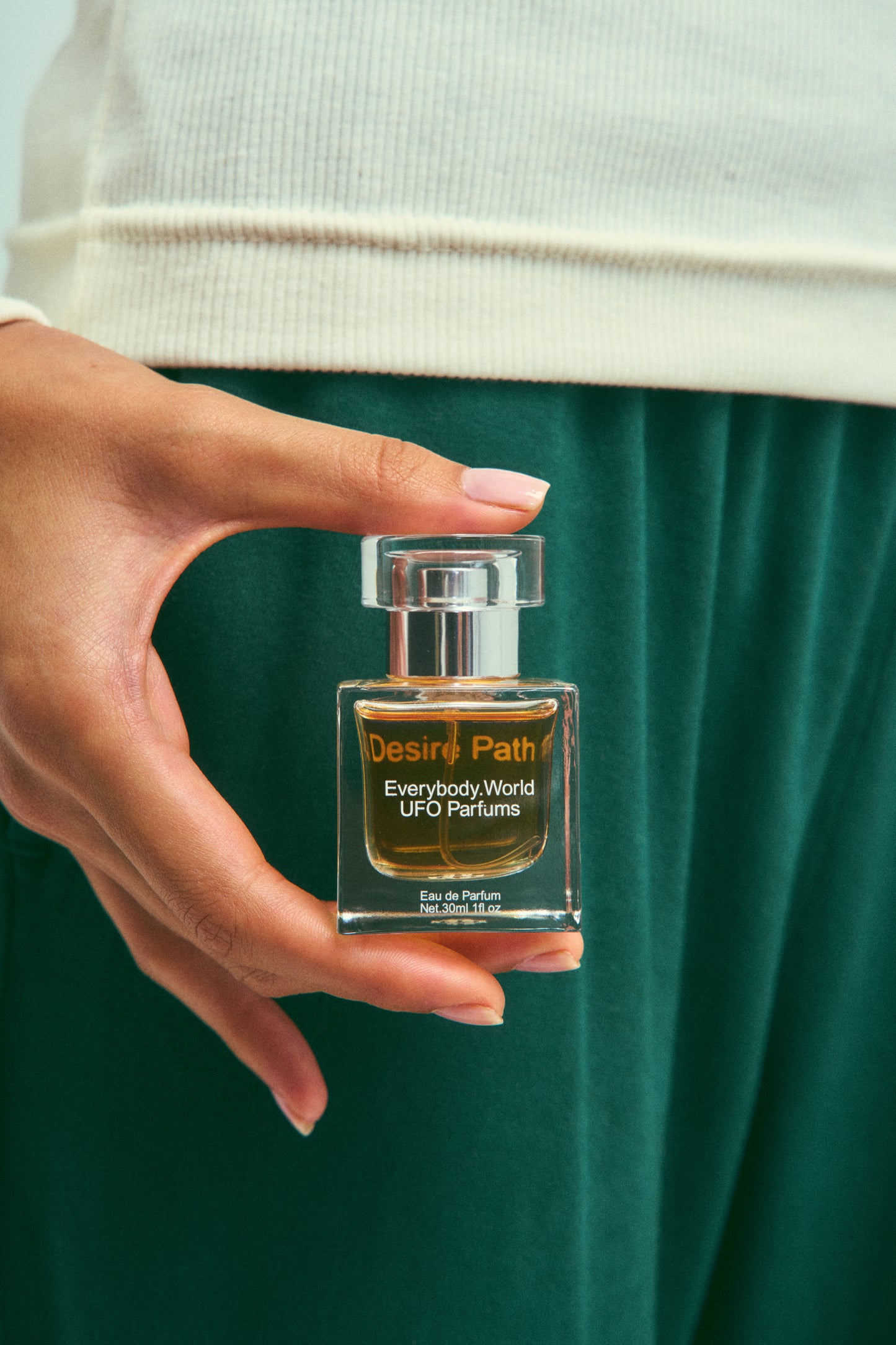 A clear glass bottle of Desire Path Eau de Parfum with a silver cap, containing a yellow-colored liquid held in a person's hand for scale.