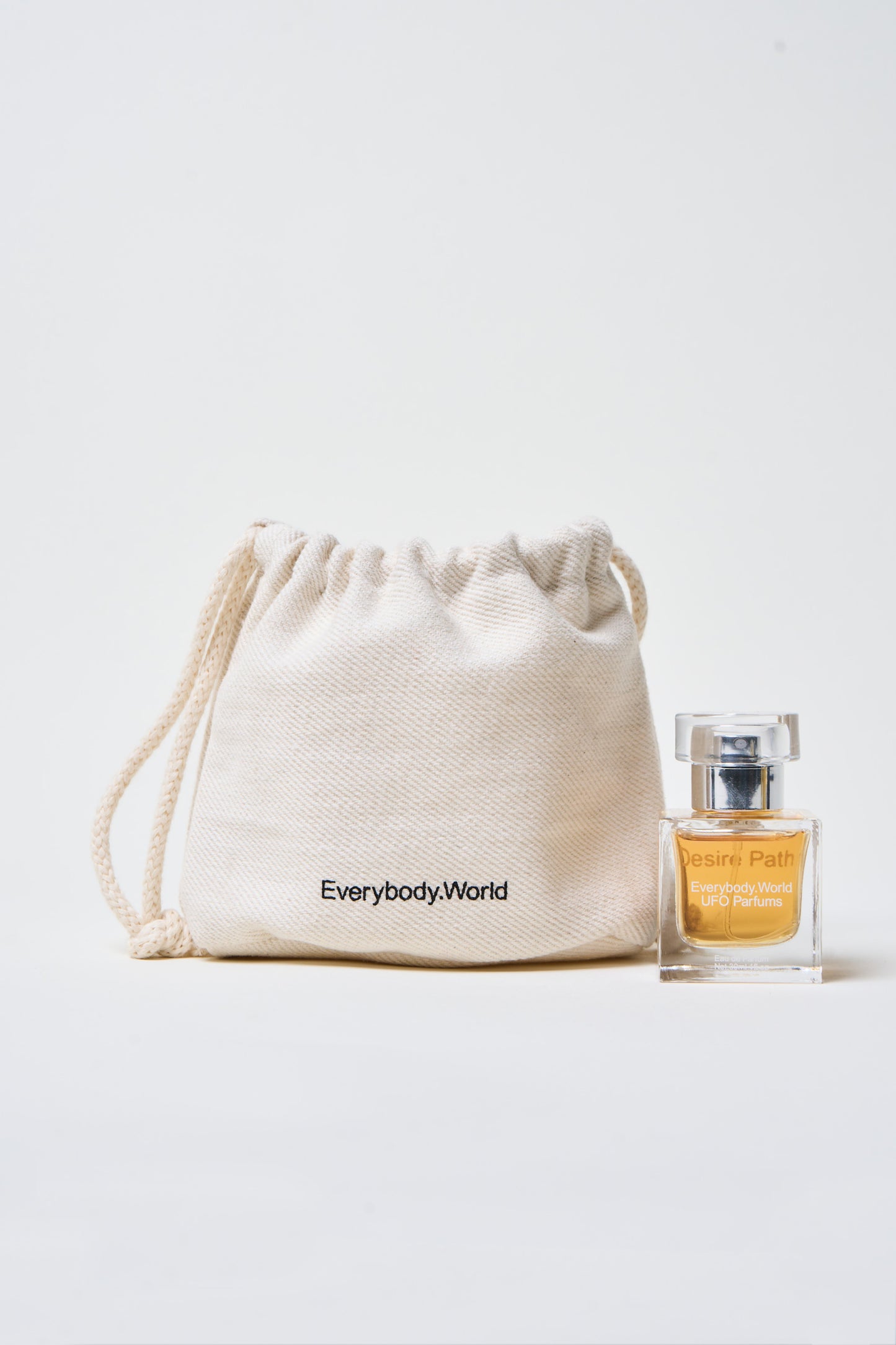 A bottle of Desire Path Eau de Parfum next to a drawstring heavy canvas bag with "Everybody.World" printed on it.