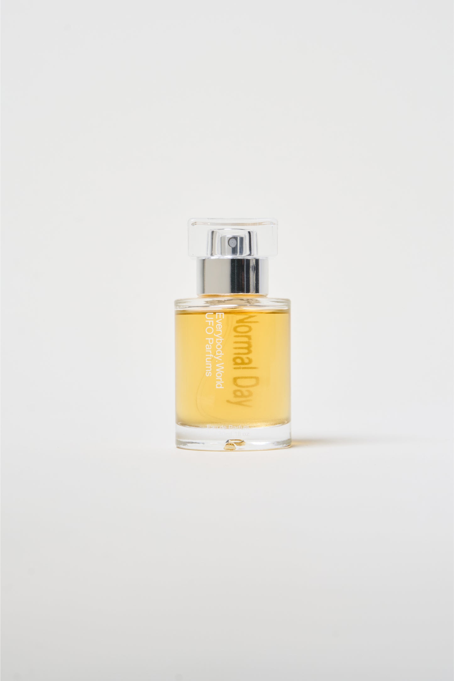 A clear glass bottle of Normal Day Eau de Parfum with label printed on the bottle, yellow-colored liquid, and a silver cap.
