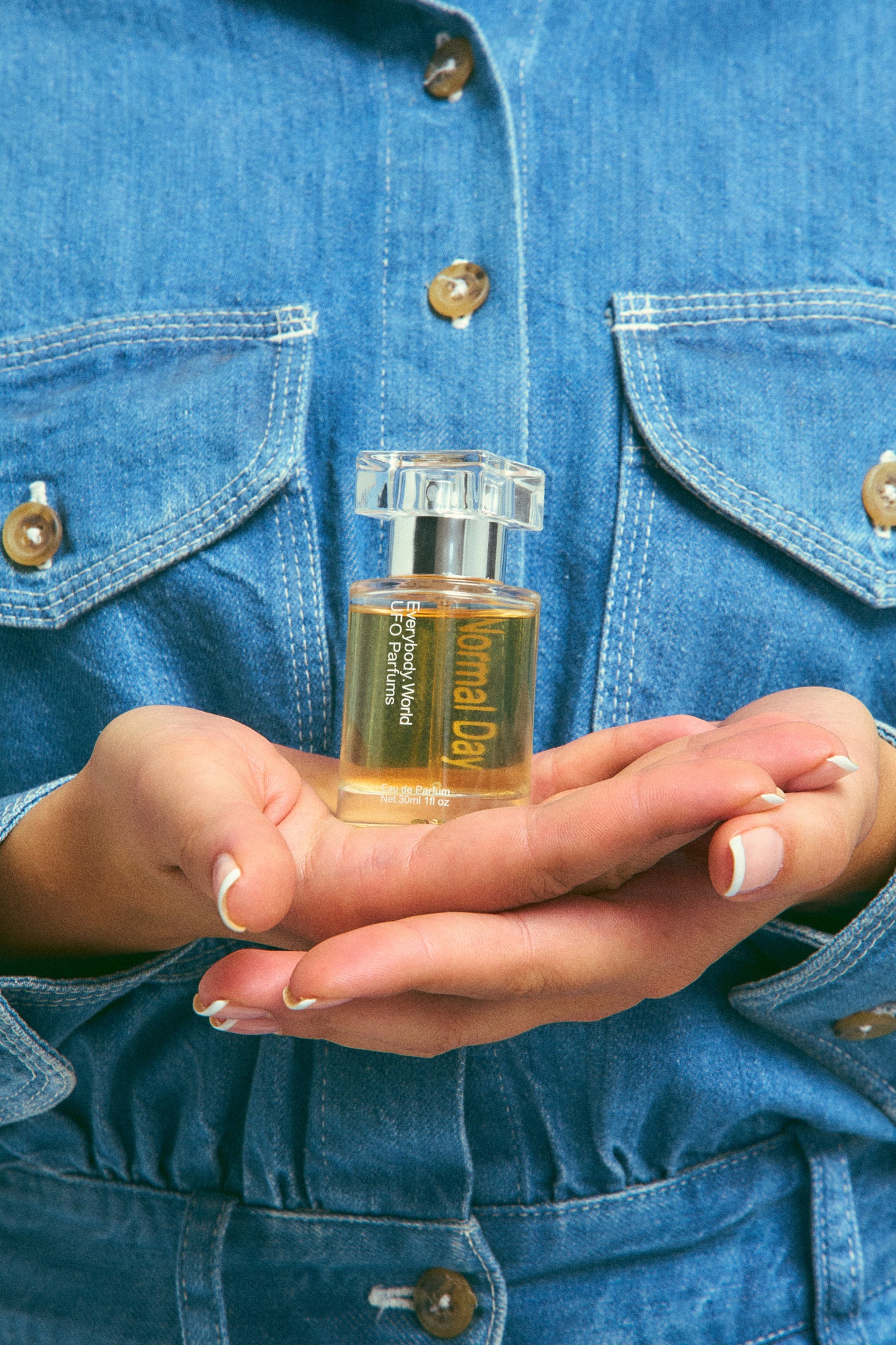 A clear glass bottle of Normal Day Eau de Parfum with a silver cap, containing a yellow-colored liquid, held in a person's palm to show scale.