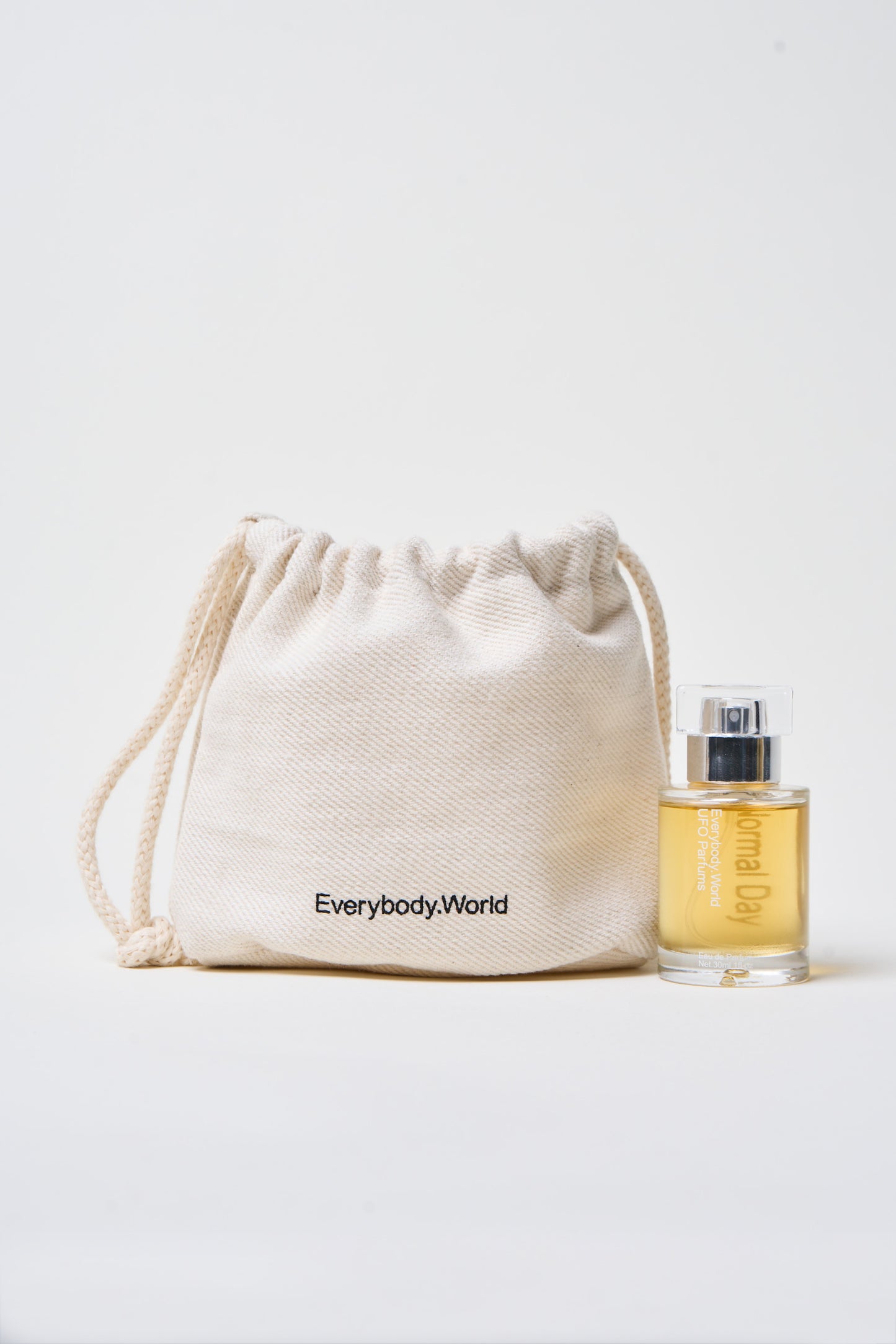 A bottle of Normal Day Eau de Parfum next to a drawstring heavy canvas bag with "Everybody.World" printed on it.