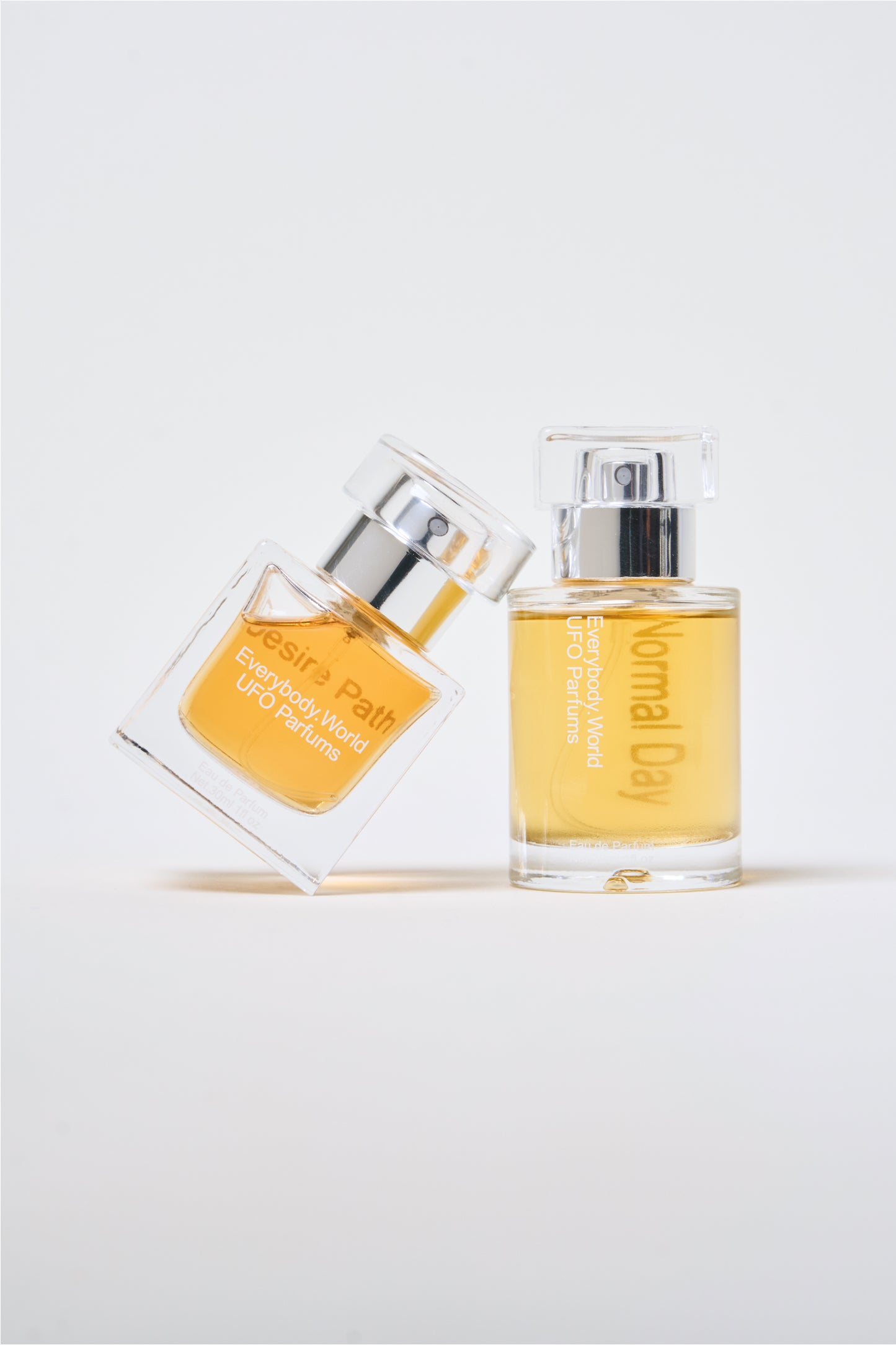 A glass bottle of Desire Path Eau de Parfum leaned against a bottle of Normal Day Eau de Parfum.