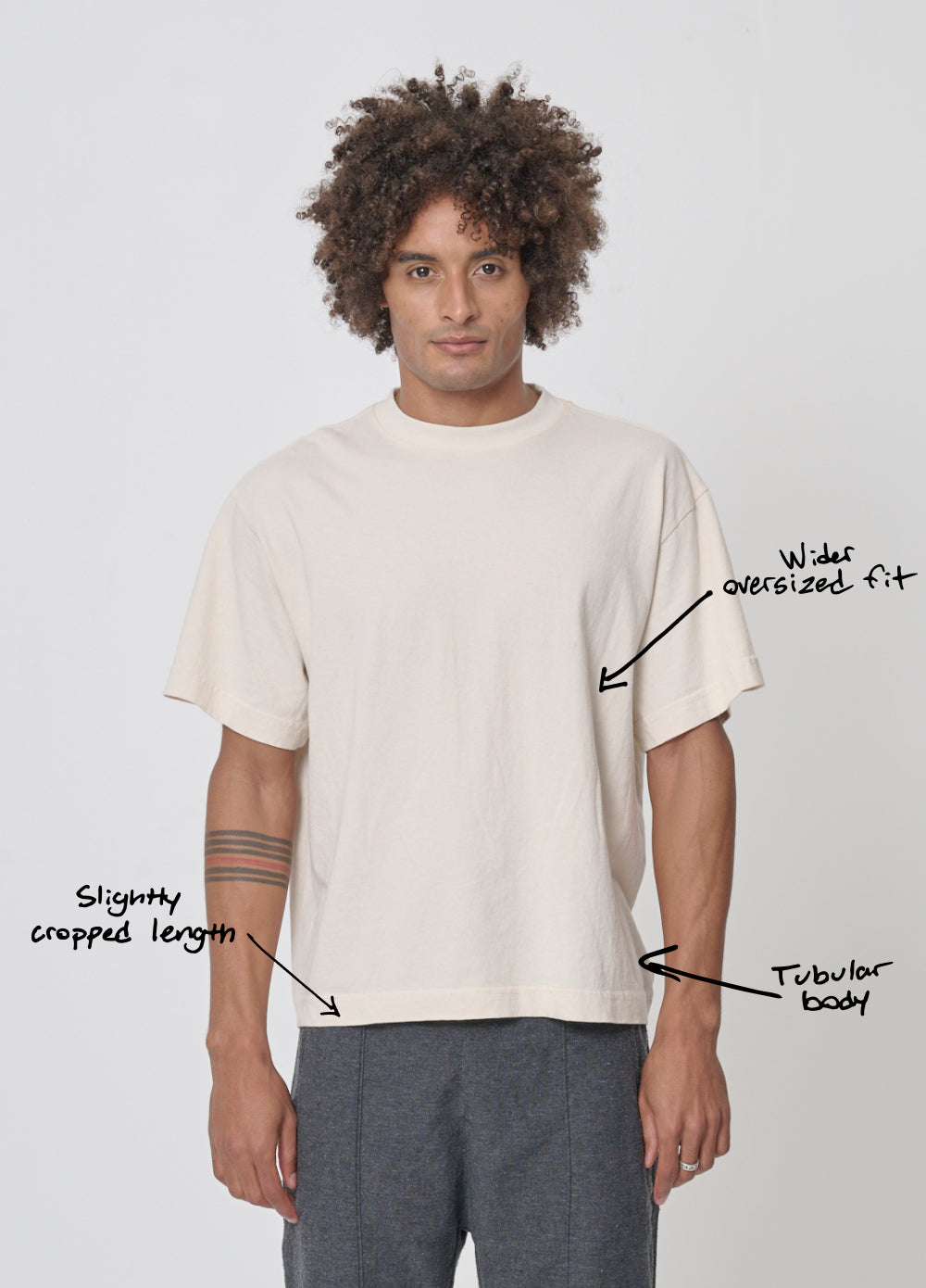 Man wearing Boxier Trash Tee. Text overlay points to the upper mid-torso and says "wider relaxed fit," lower on the side of the shirt says "tubular body” and at the hem, "Slightly cropped length."