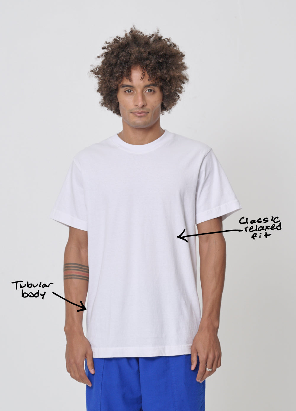 A man in a Short Sleeve Trash Tee. Annotation points to the upper mid-torso and says "classic relaxed fit" and lower on the side of the shirt says "tubular body.”