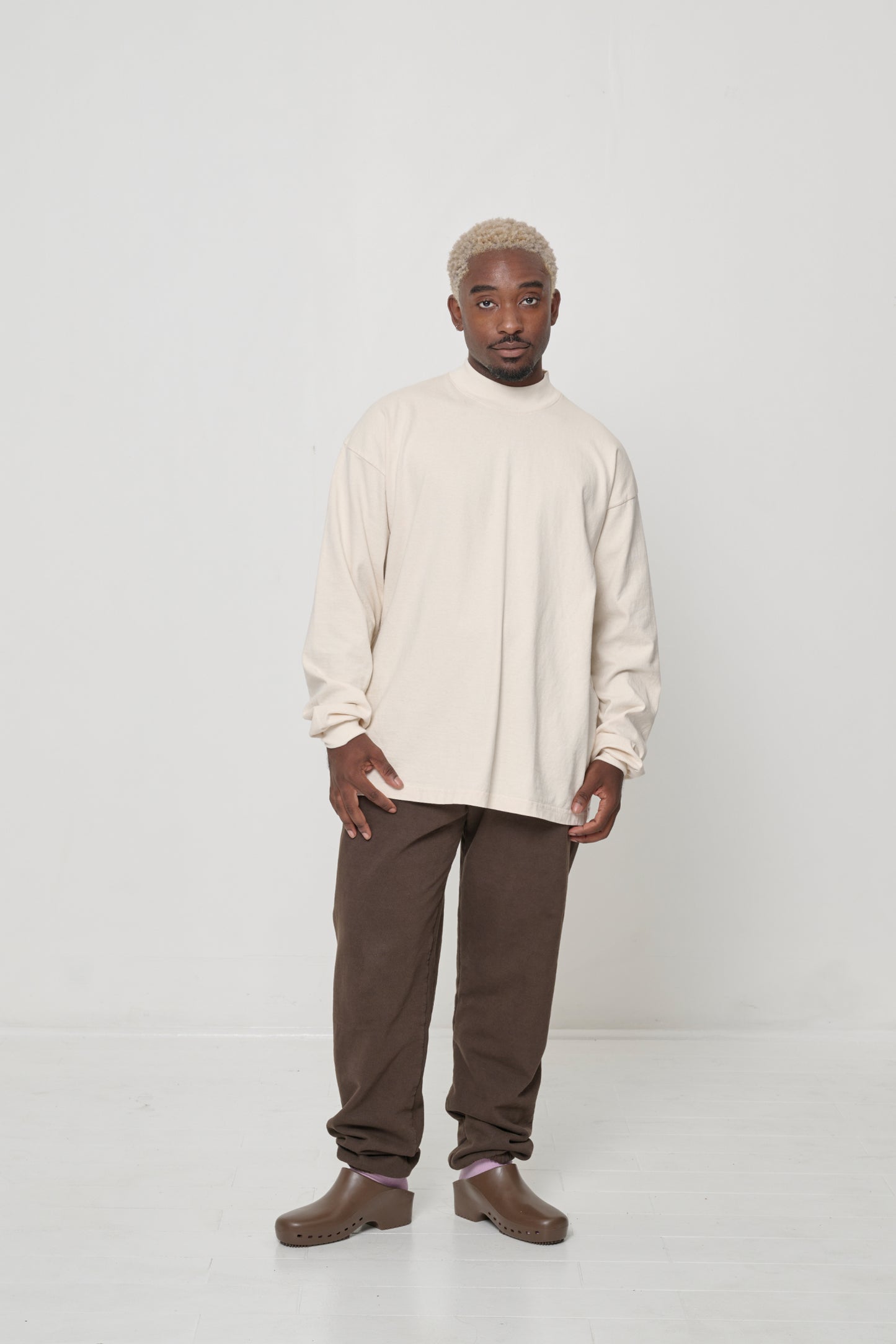 Full length front view of person wearing a Scour (pale beige) JAS Mockneck long-sleeve t-shirt, paired with brown Trash Sweatpants and Brown Classic Calzuro Clogs.