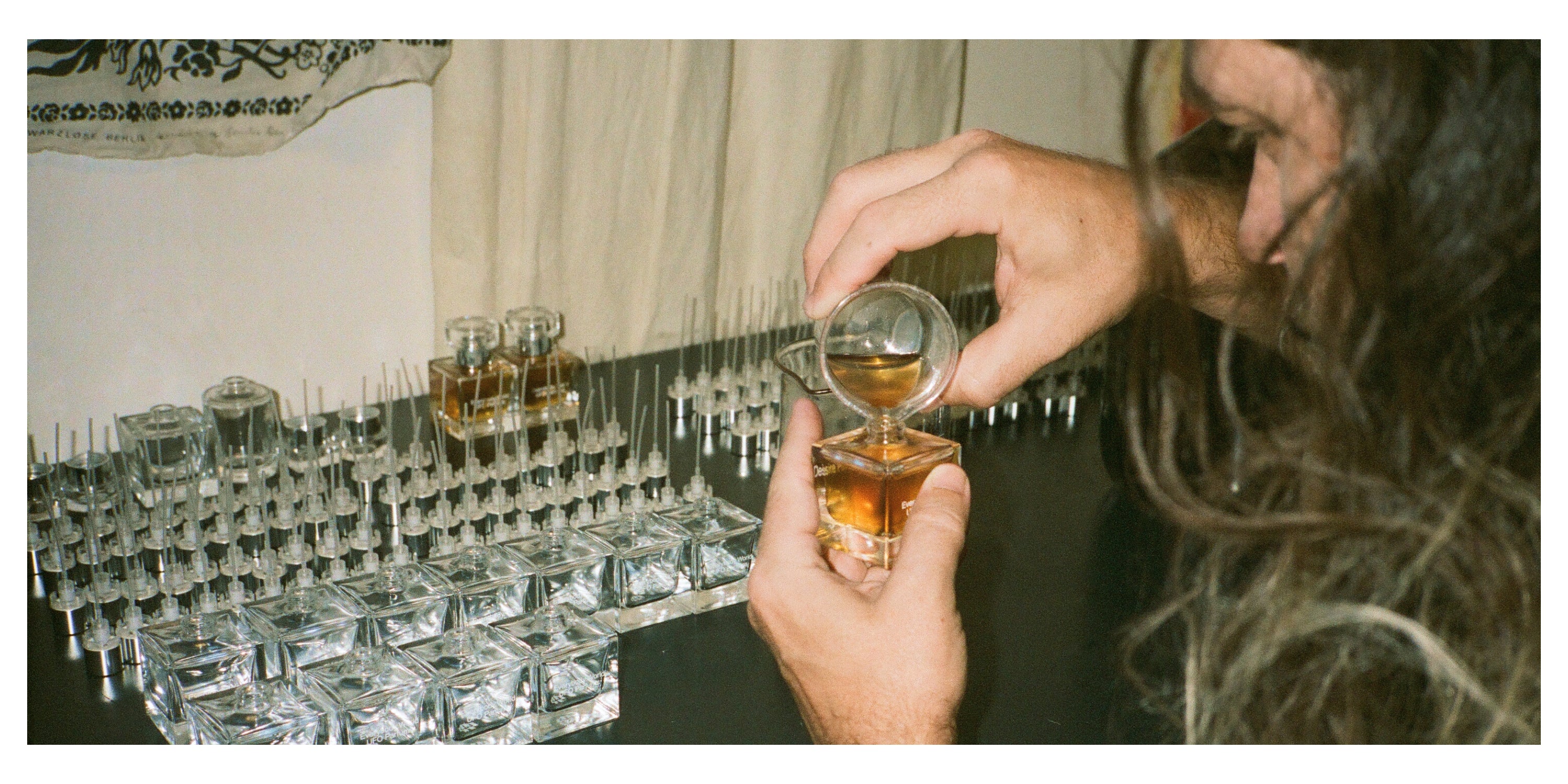 Hands pouring perfume into a Desire Path bottle. Top portion of pourer's face is in frame. Rows of empty bottles and nozzles in background.