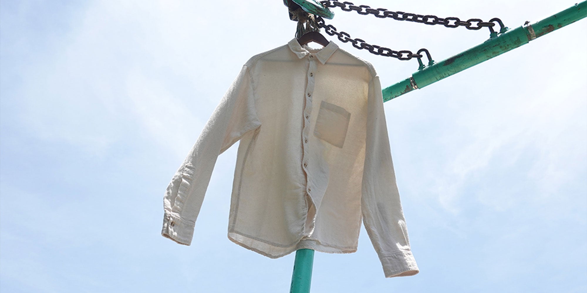 Light beige button up shirt on a hanger, hung on green poles and chains with clouds and blues sky in background.