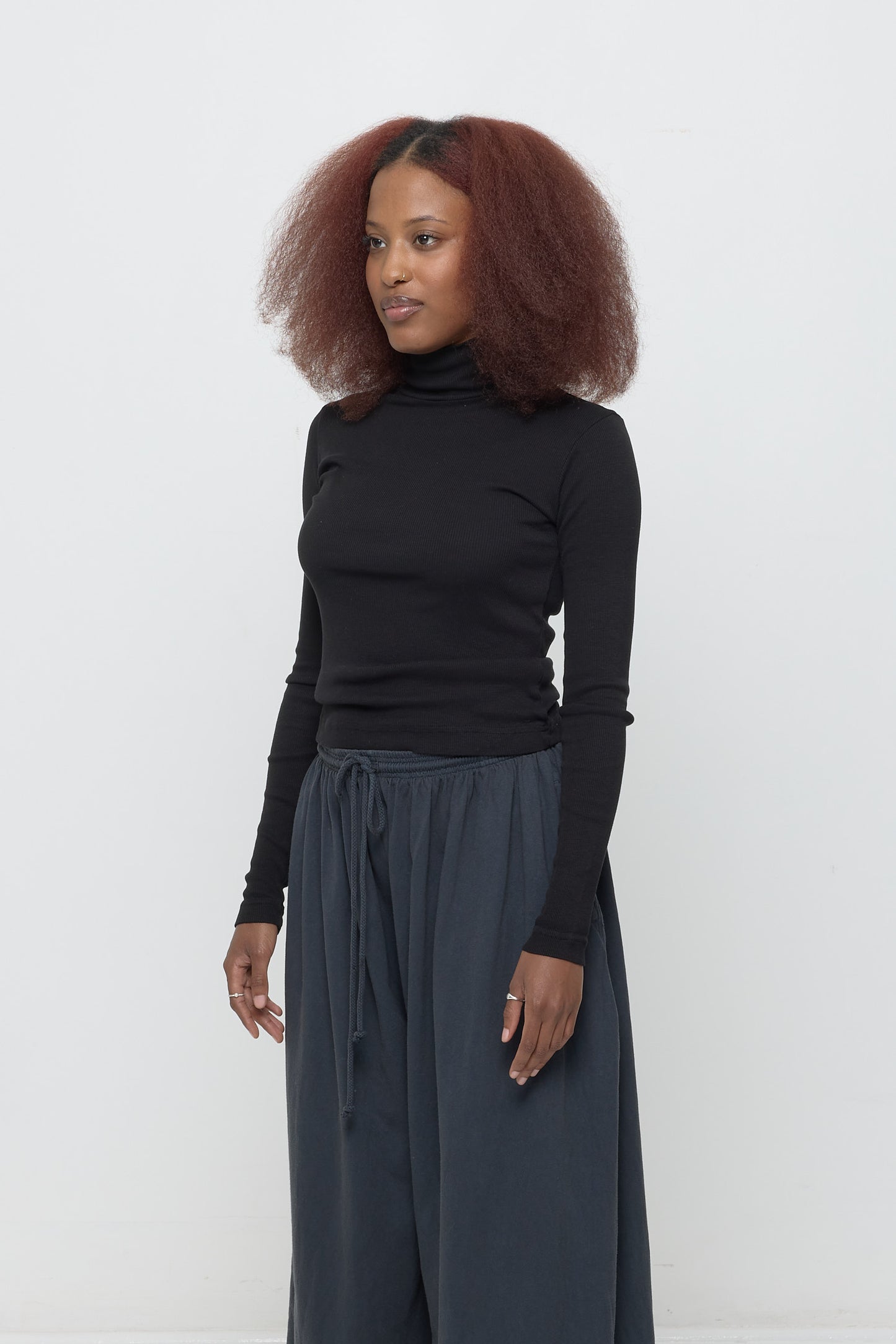 A person turned 45-degrees from the front, wearing a Rib Turtleneck in Black, showing the side seams, heavy hem, and fitted silhouette.