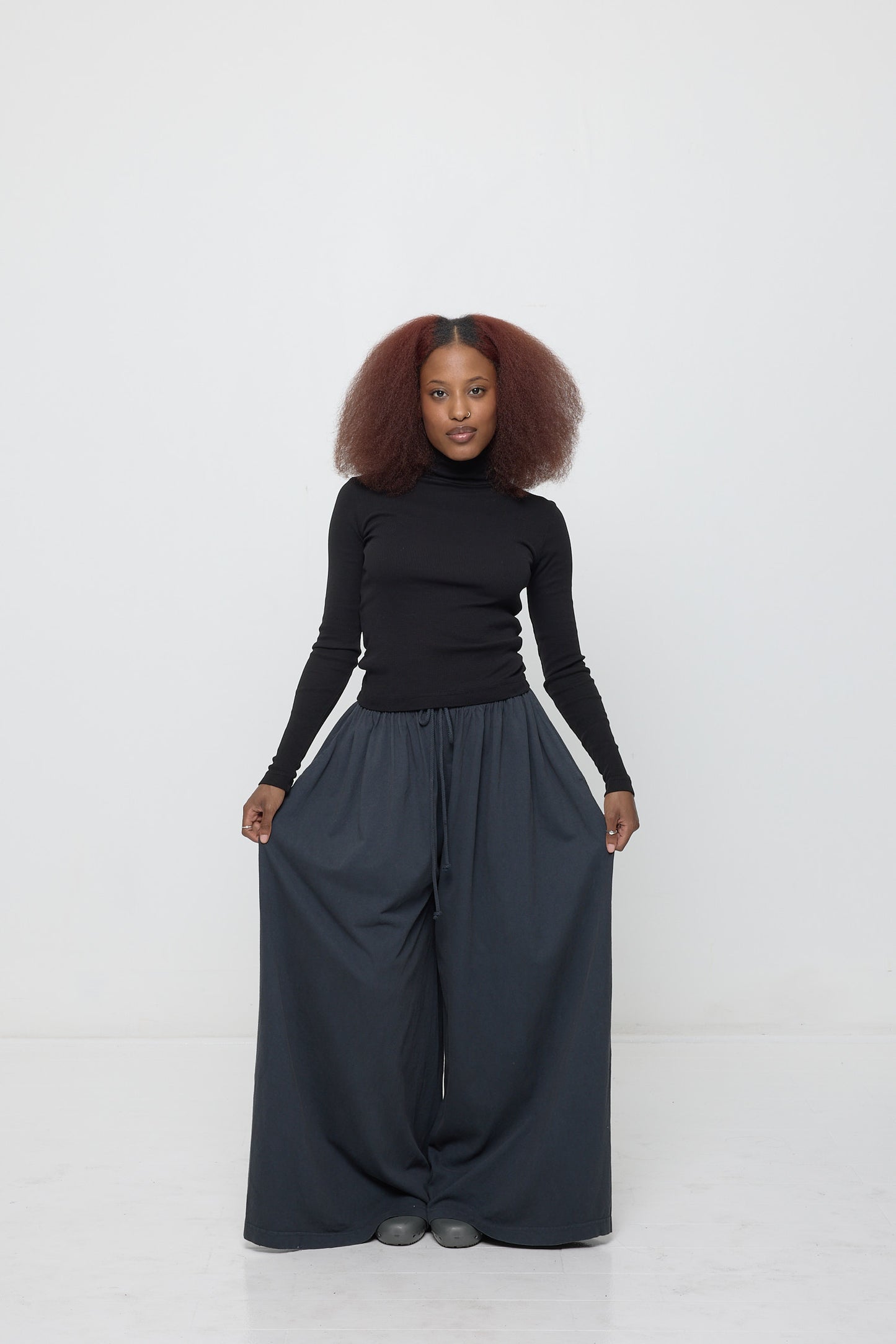 A woman wearing a Black Rib Turtleneck, holding the sides of her Tubular Pants, focusing attention on the fitted sleeves.
