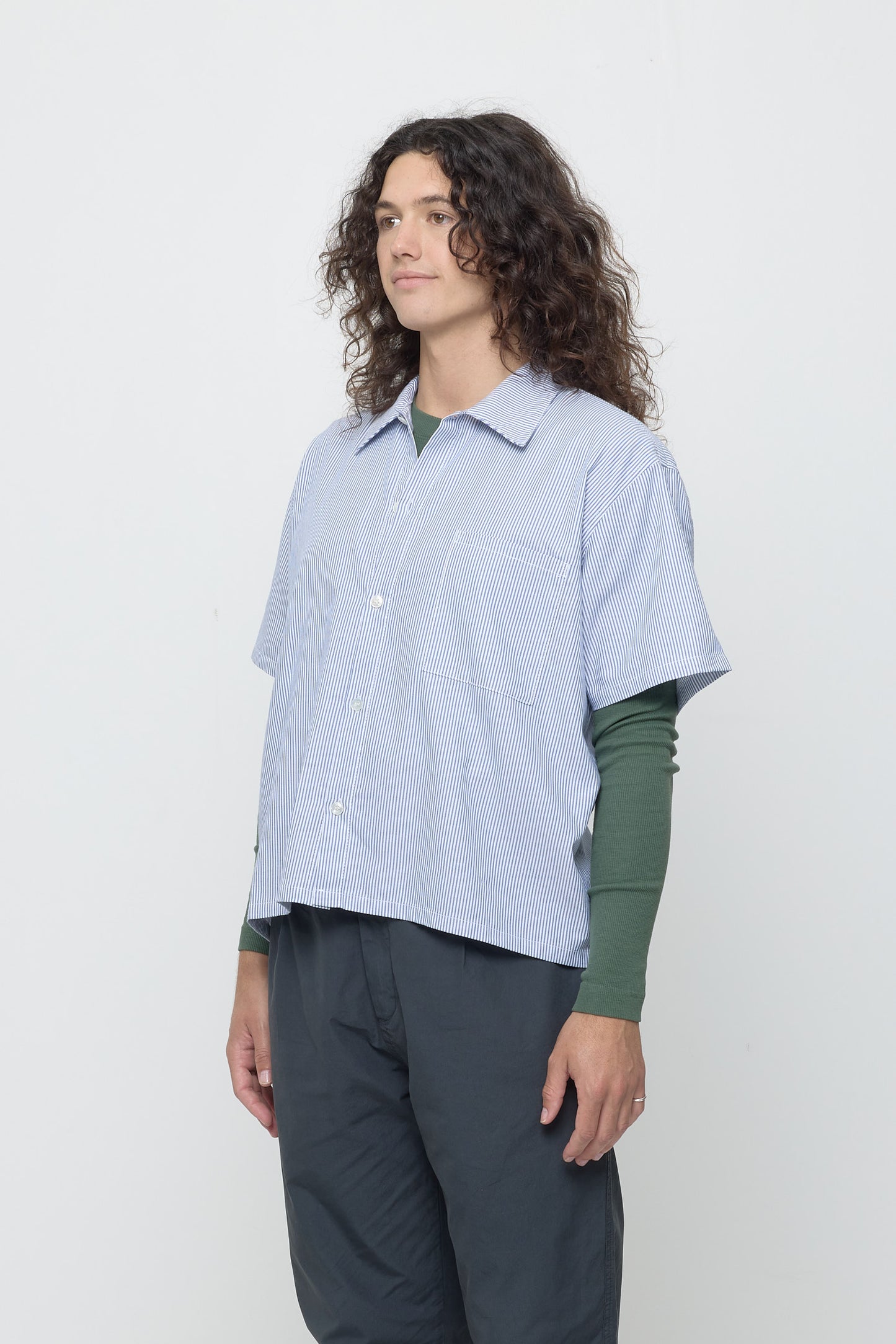 A person in a blue Stripe Poplin Button Up Shirt in a ¾ front view, highlighting the shirt's loose sleeves and chest pocket.