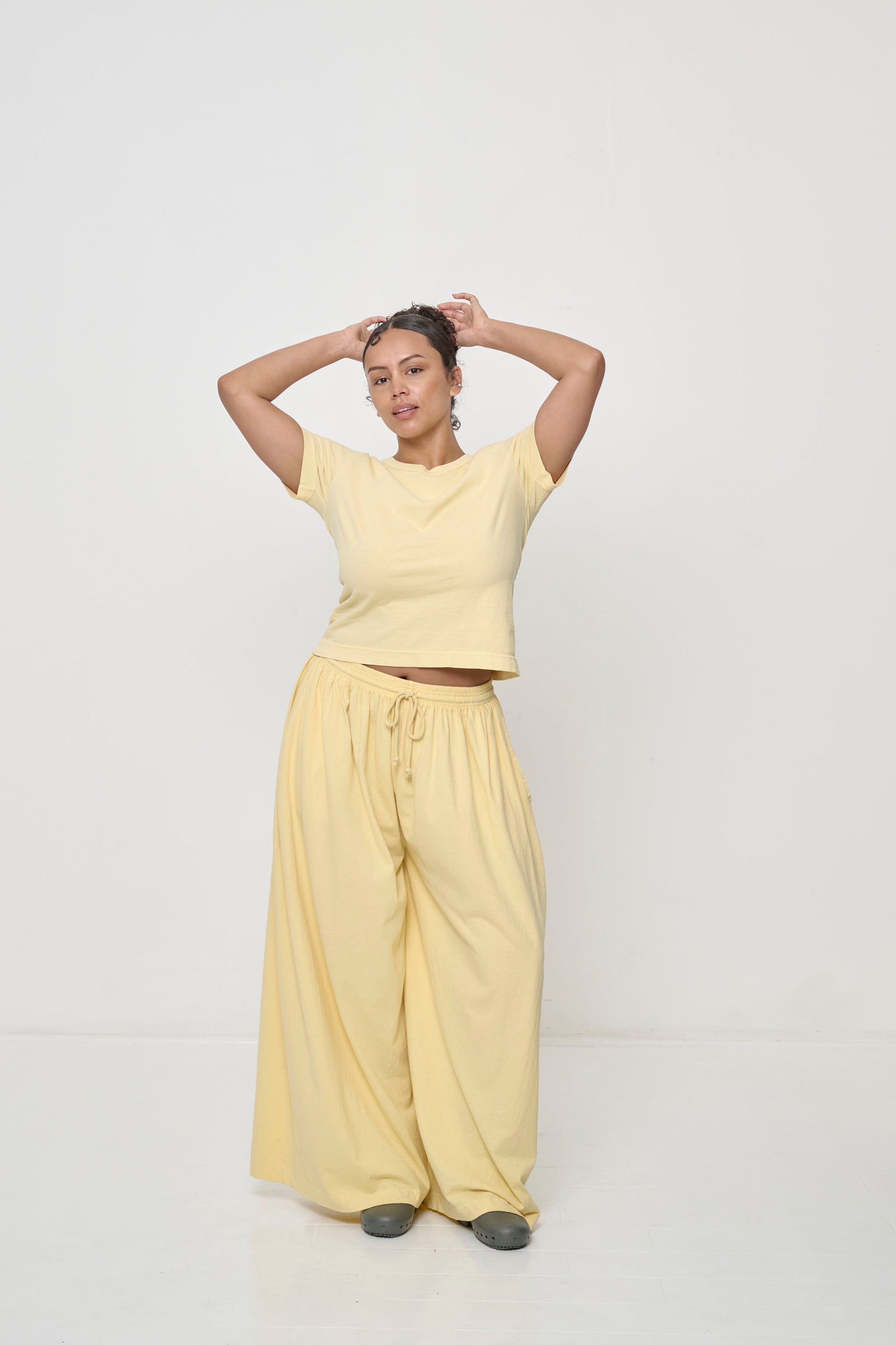Woman in French Butter Tailored Trash Tee and Apartment Pants, hands on head, shirt hem raised just above navel.