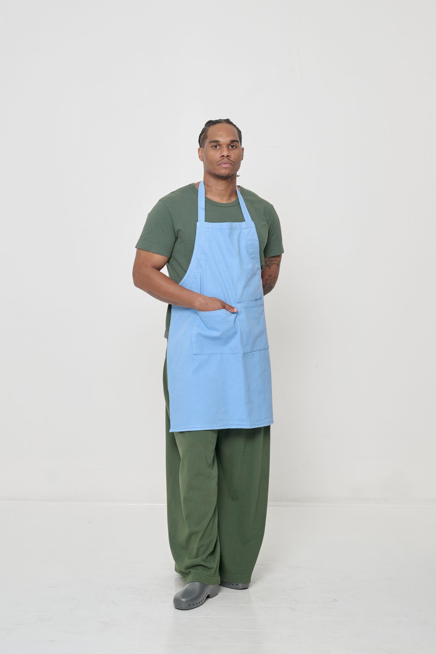 Man in light blue apron, Pine Tailored Trash Tee, and Apartment Pants, hand in pocket, displaying shirt's slim sleeves.
