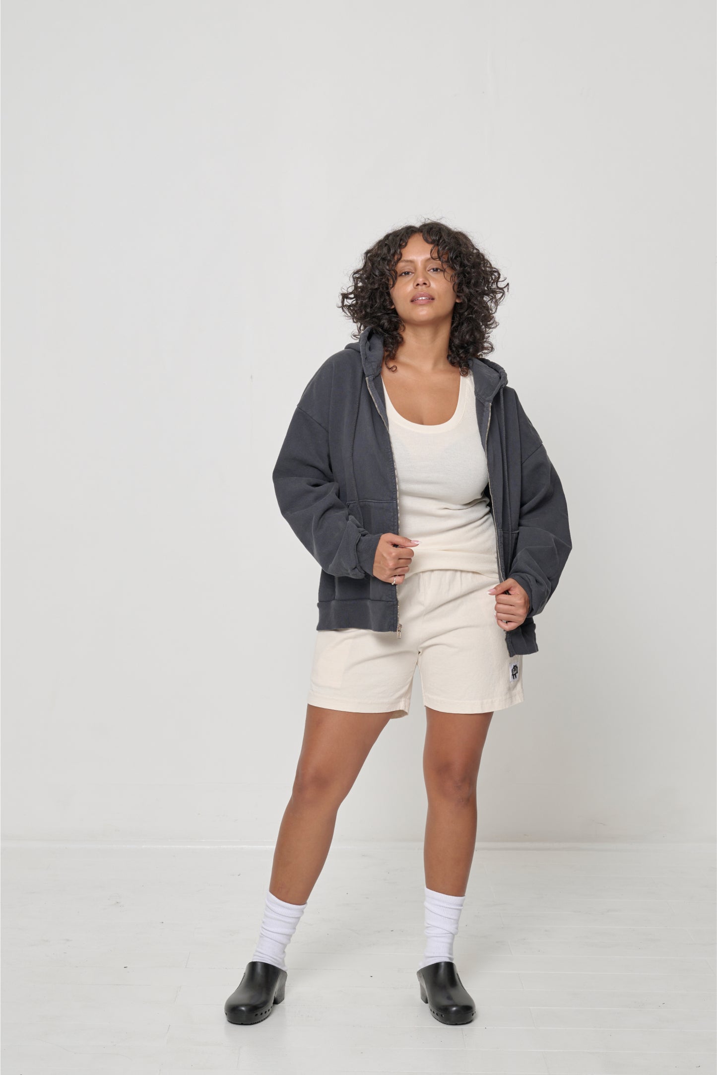 Woman in Natural The Tank, layered under a Slate Trash Zip Up Hoodie, and Scour Everyday Shorts.