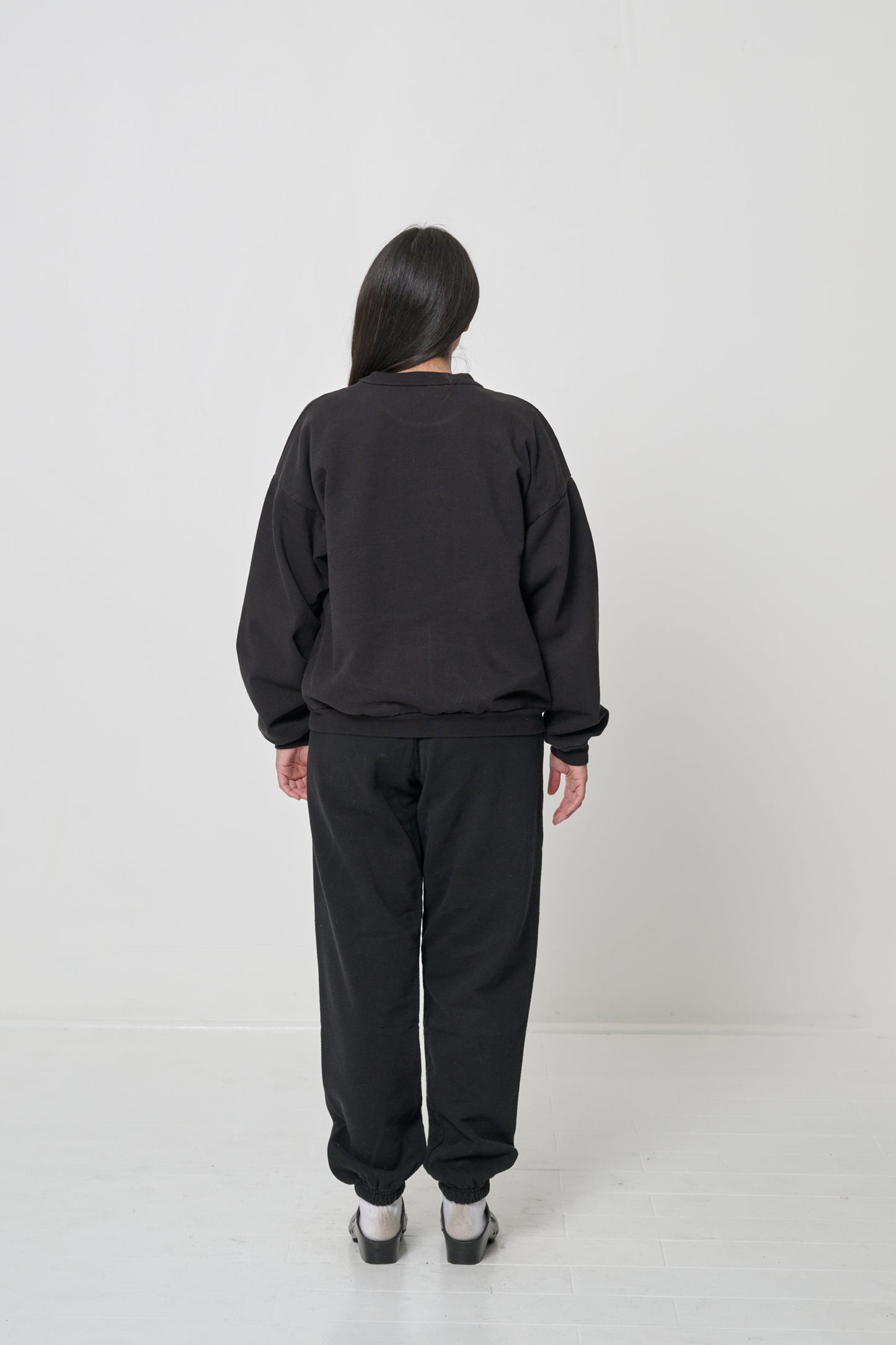 Woman facing straight back in a Black Trash Crewneck. Relaxed fit throughout until the rib hem which is fitted around the hips.
