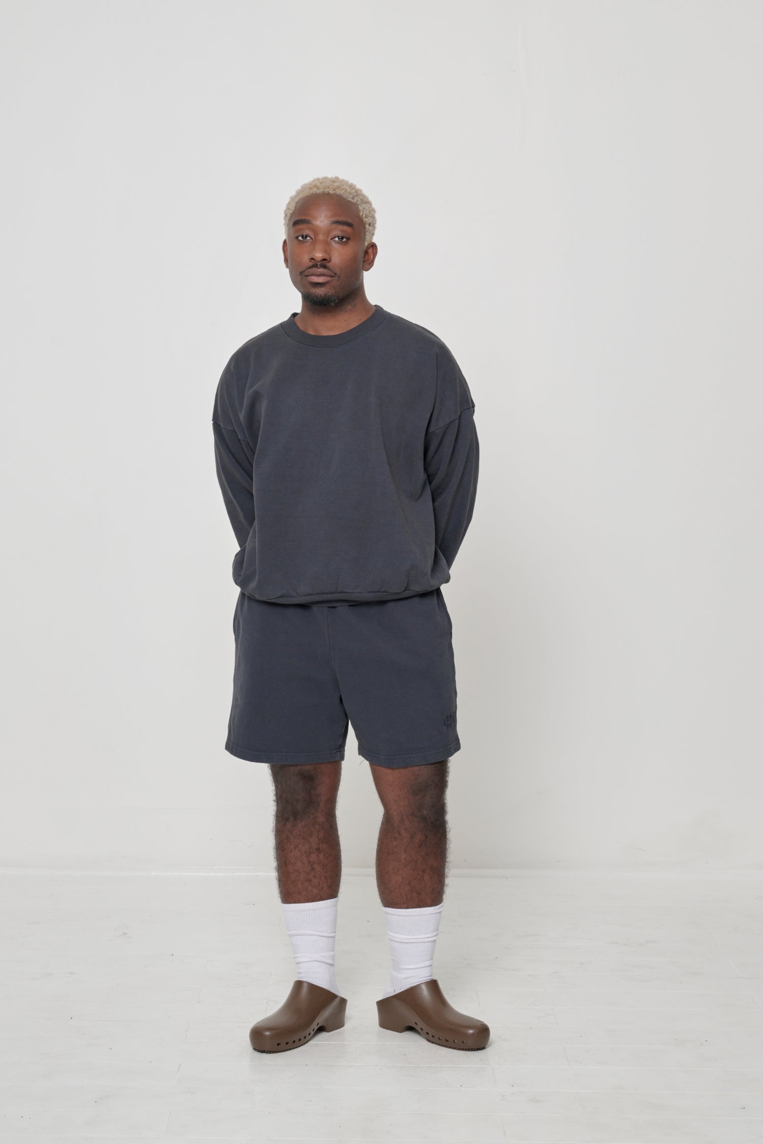 Man in matching Slate Trash Crewneck and Trash Fleece Everyday Shorts, with White Squishy Socks and Brown Calzuro Classic Clogs.