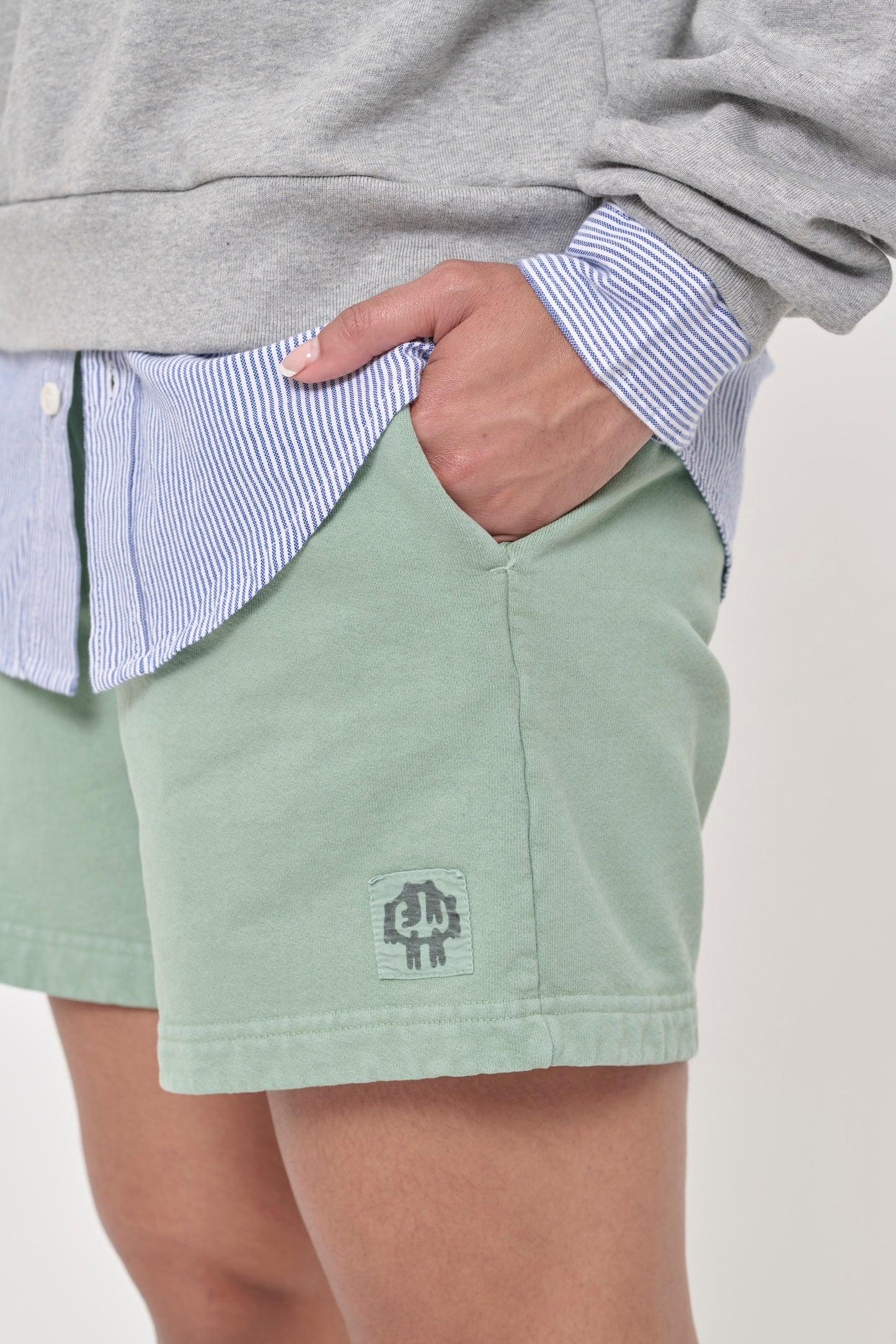 Closeup of hand in left side pocket of Wasabi Trash Fleece Everyday Shorts, shows printed E.W logo patch on bottom left leg.