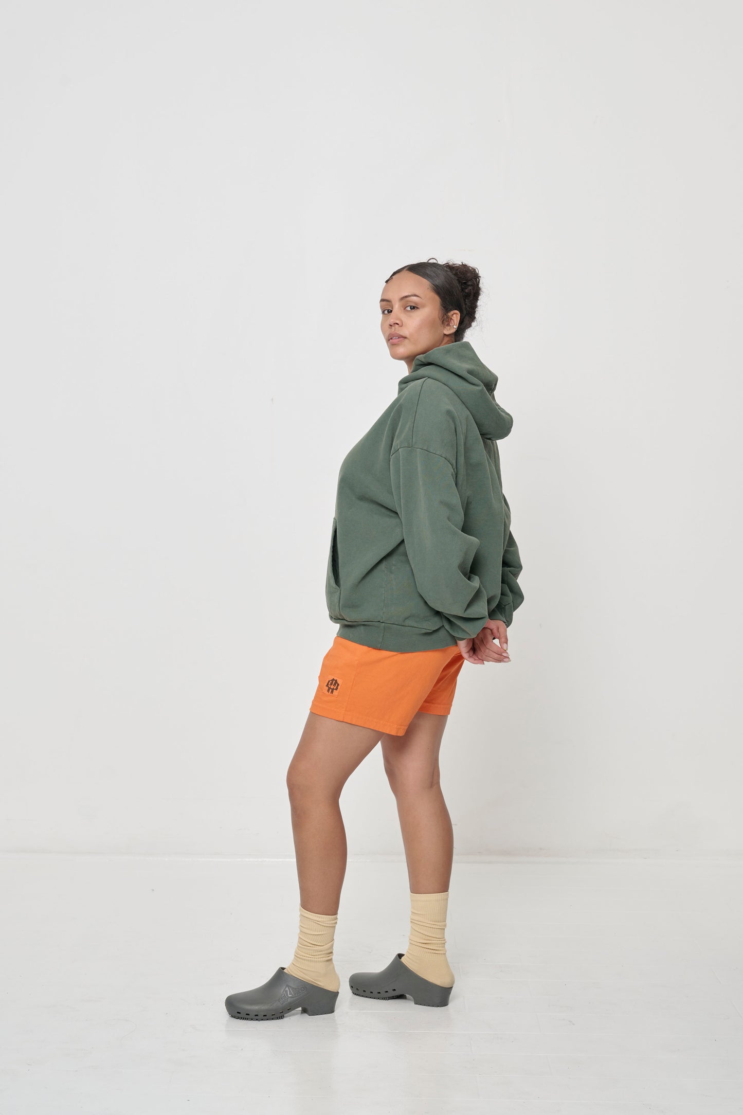 Profile full length view: woman wearing a Pine Trash Hoodie with hands behind her back, showing the shoulder and side seams.