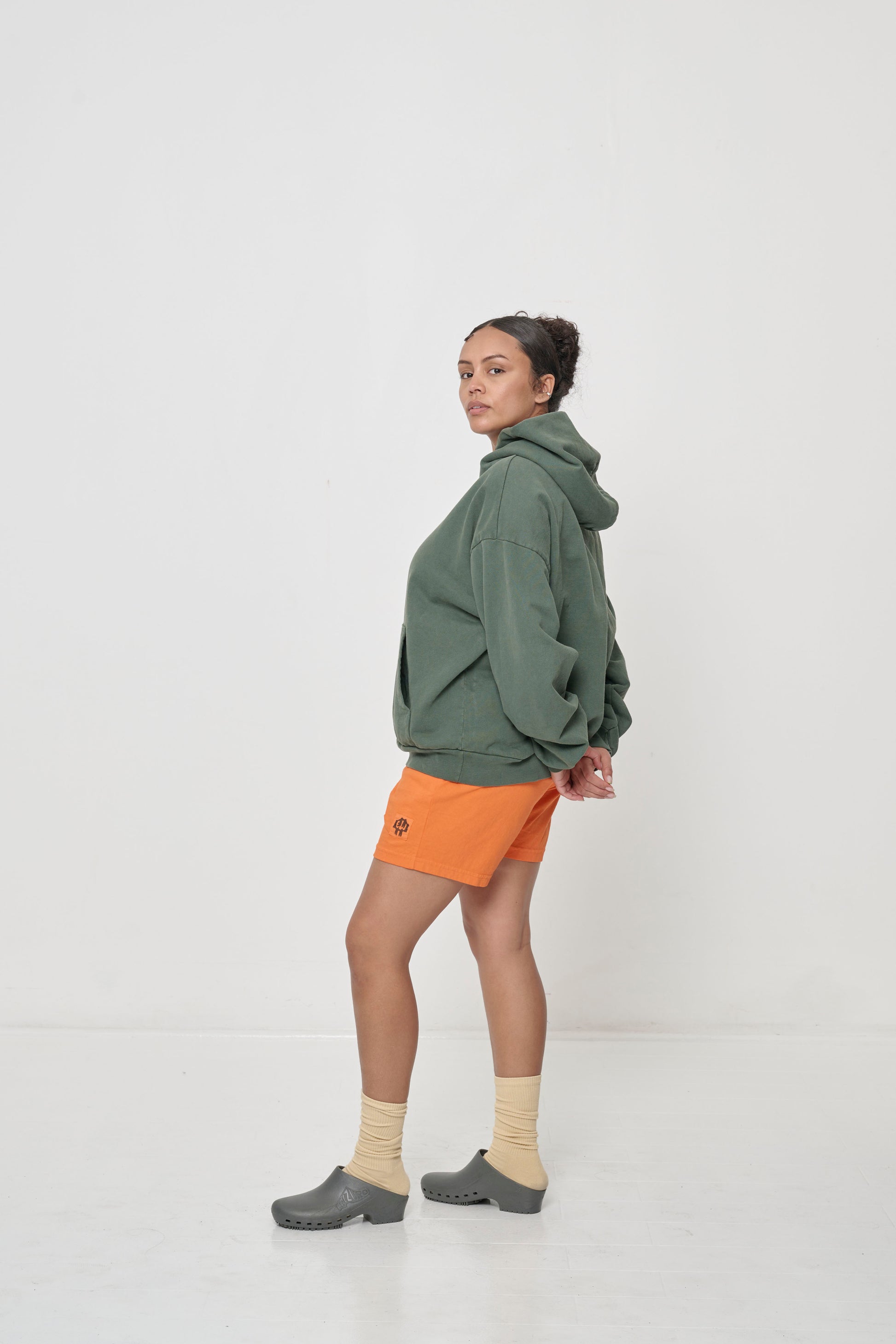 Profile full length view: woman wearing a Pine Trash Hoodie with hands behind her back, showing the shoulder and side seams.