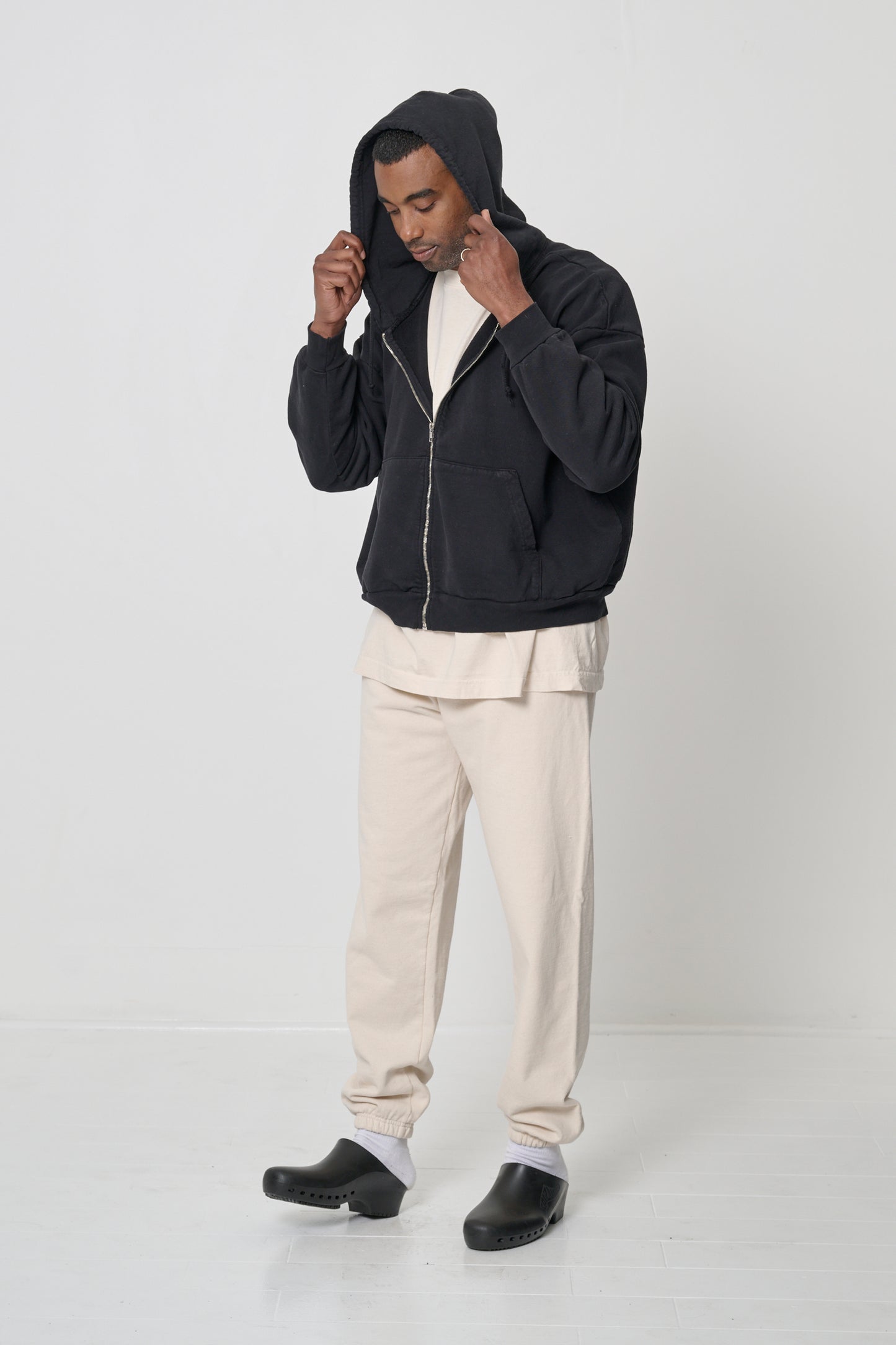 Man facing 45-degrees, wearing a Black Trash Zip Up Hoodie, adjusting hood, which he's worn on his head.