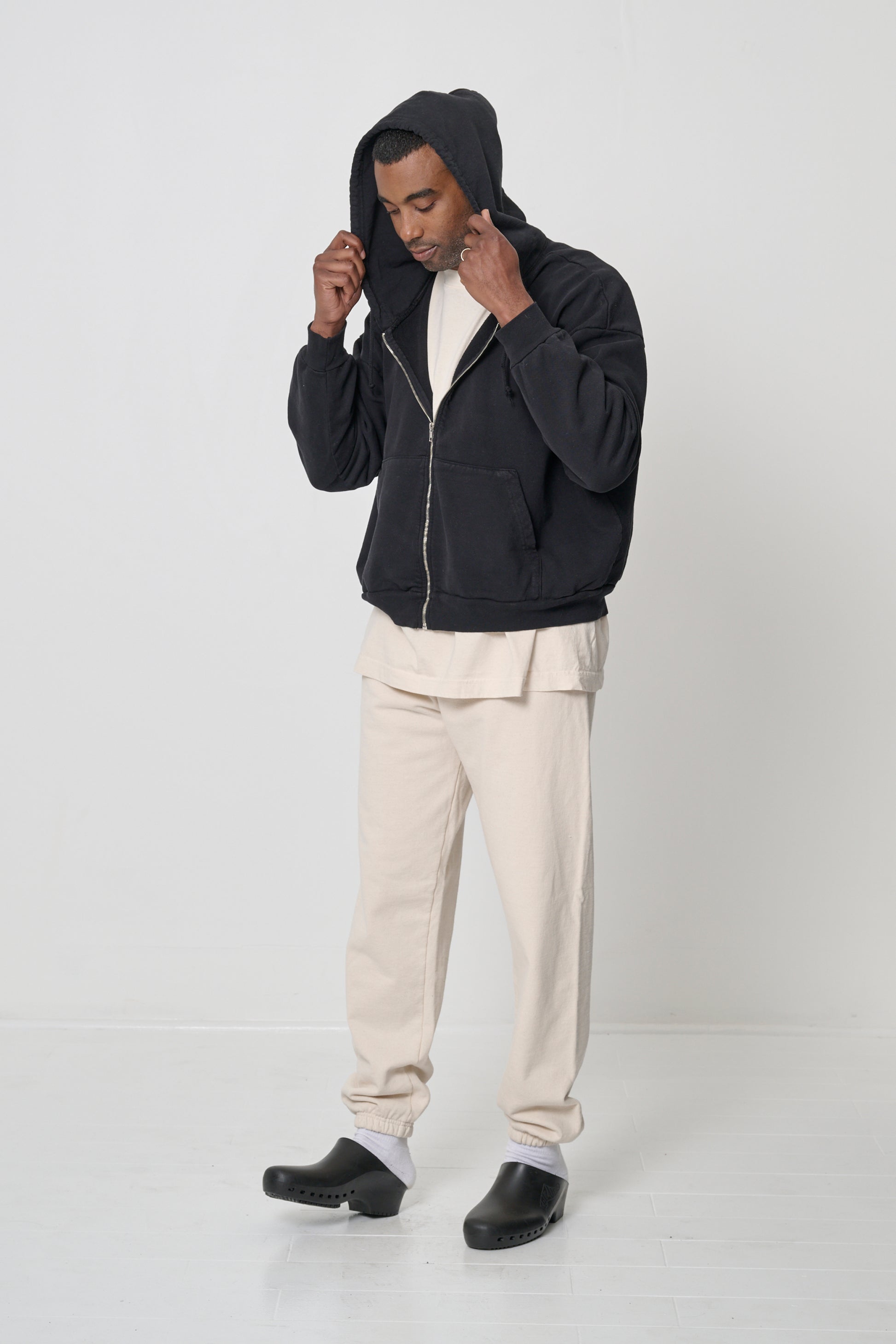 Man facing 45-degrees, wearing a Black Trash Zip Up Hoodie, adjusting hood, which he's worn on his head.