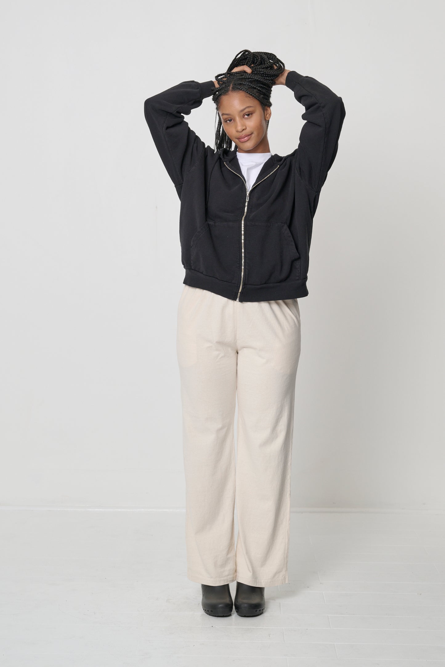 Front view: woman wearing a zipped Black Trash Zip Up Hoodie with hands atop her head, raising the hem to her upper hip.