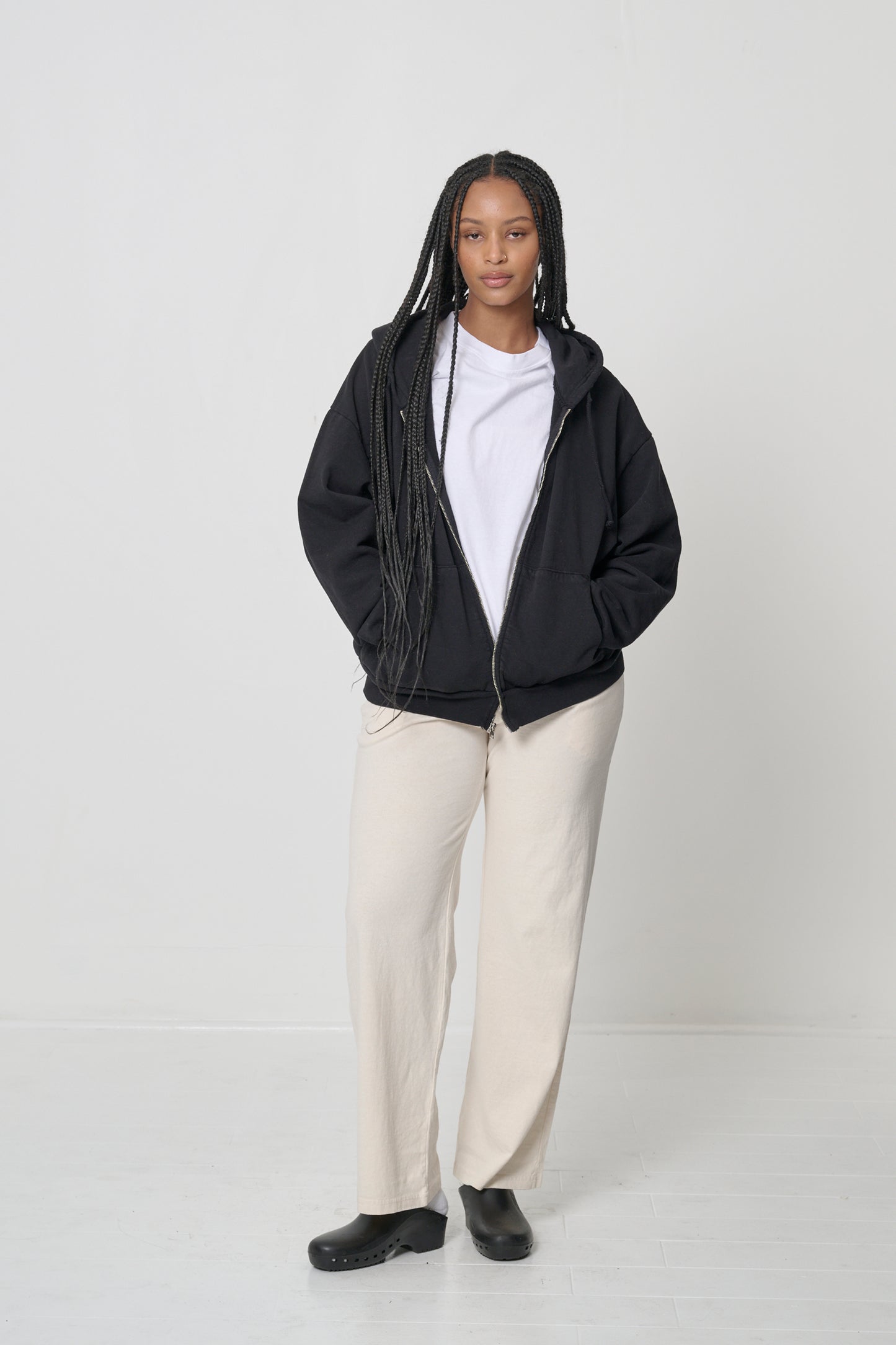 Front view: woman wearing an unzipped Black Trash Zip Up Hoodie with hands in the kangaroo pockets.