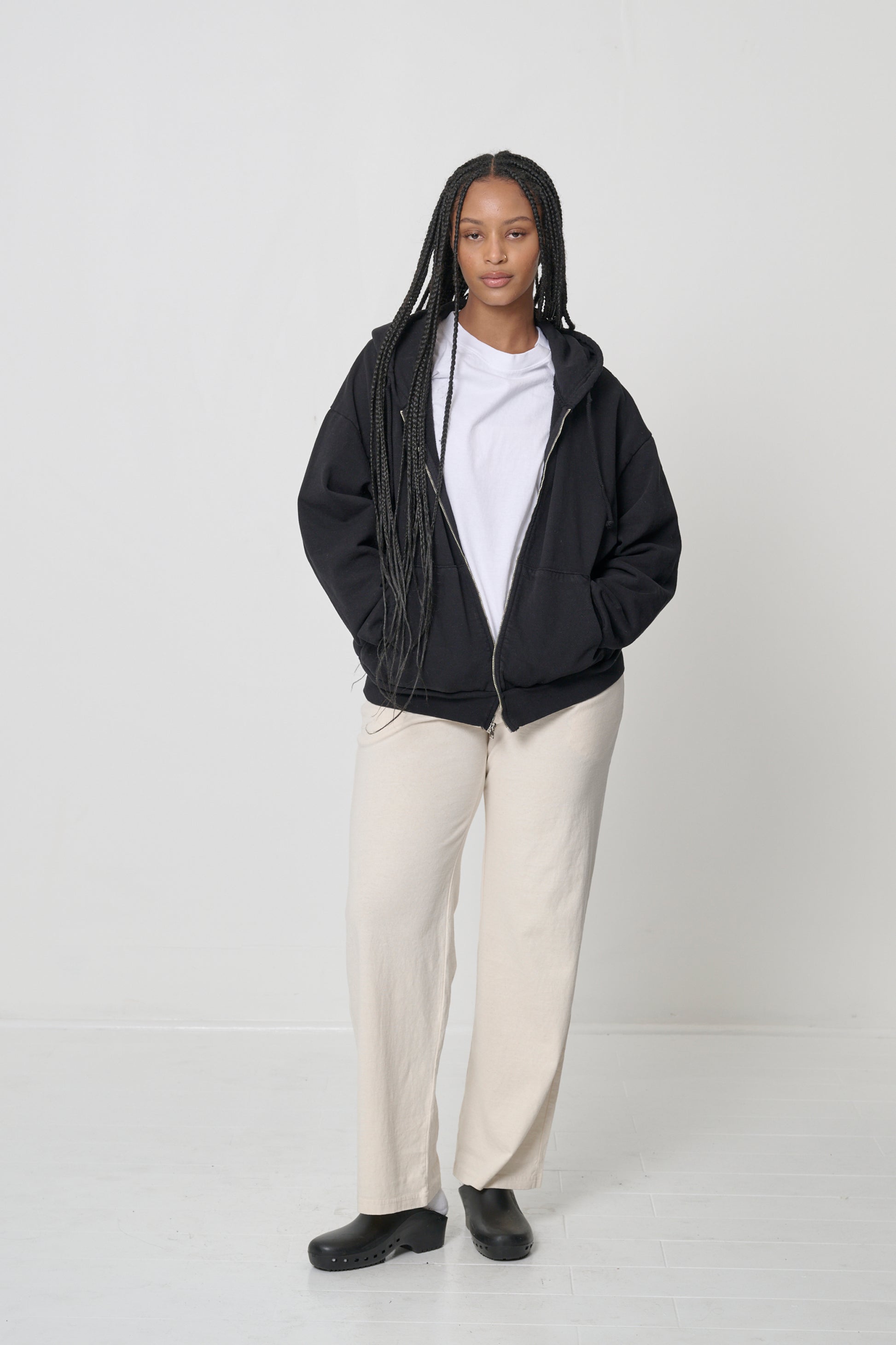 Front view: woman wearing an unzipped Black Trash Zip Up Hoodie with hands in the kangaroo pockets.